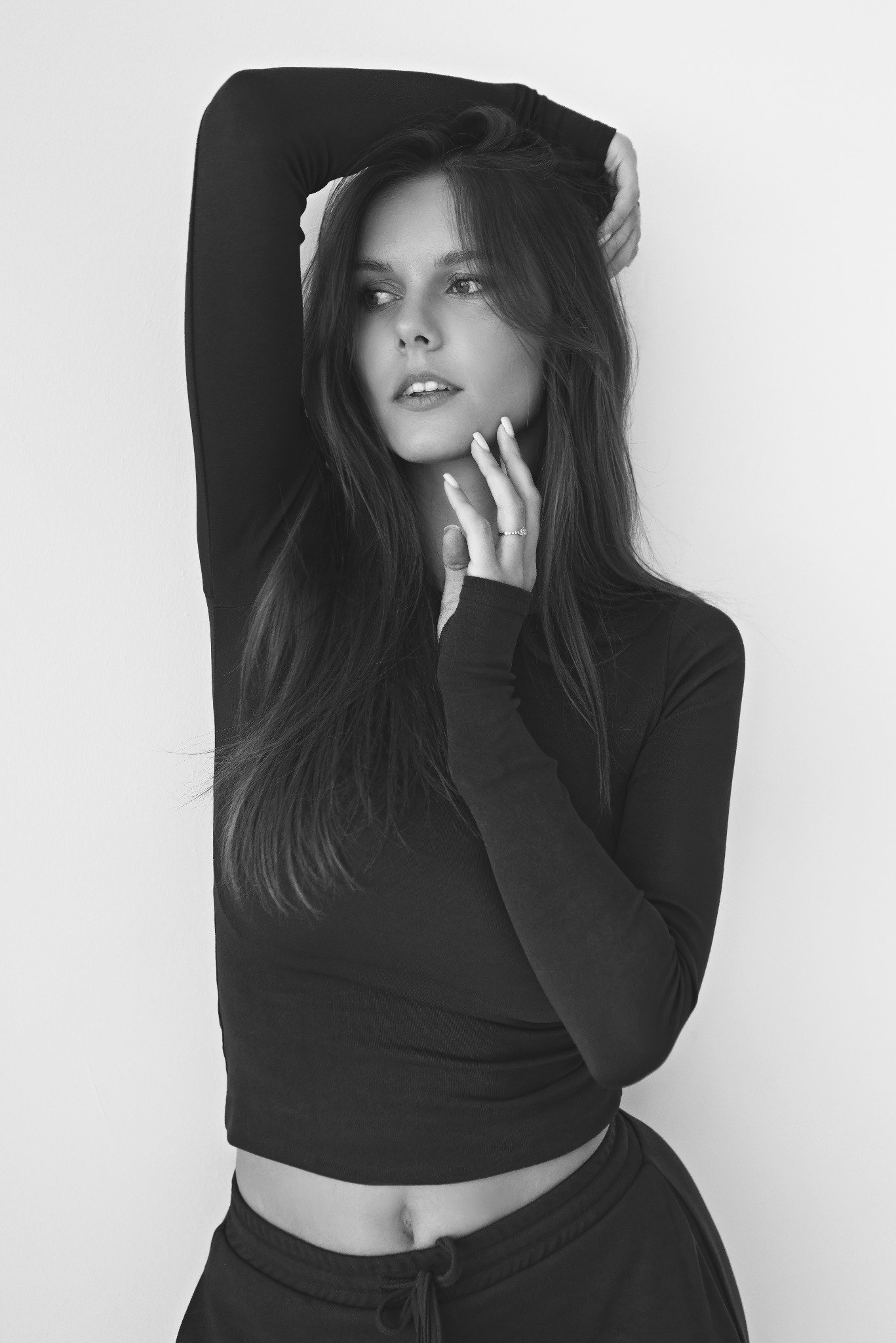 Czarno-białe portretowe zdjęcie młodej kobiety z długimi włosami, ubranej w czarny top z długim rękawem i spodnie dresowe, pozującej z rękami uniesionymi nad głową i dłońmi delikatnie dotykającymi...