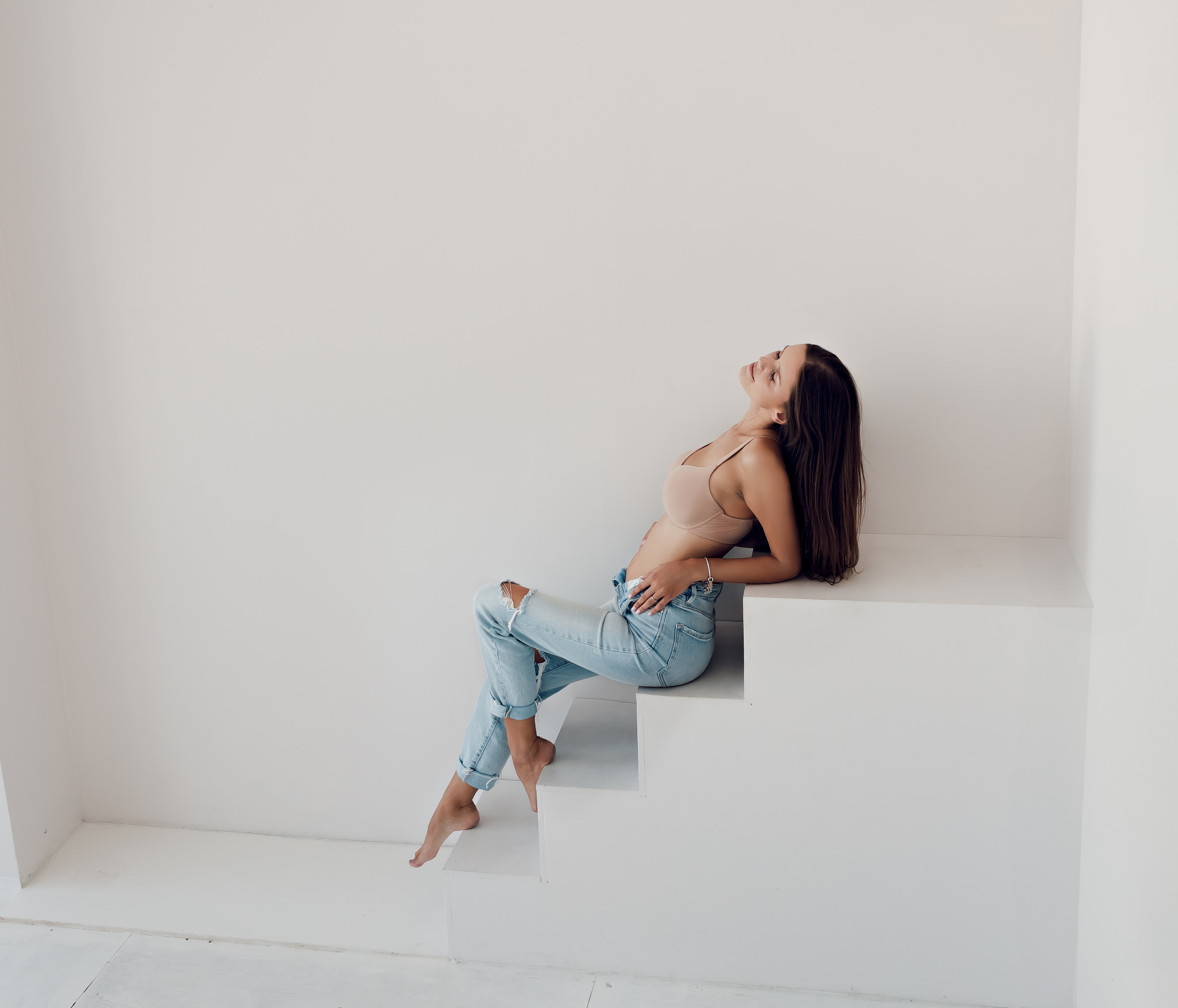 Kobieta w bieliźnie i podartych jeansach pozuje na białych schodach w minimalistycznym studio, głowa odchylona do tyłu, delikatne światło.