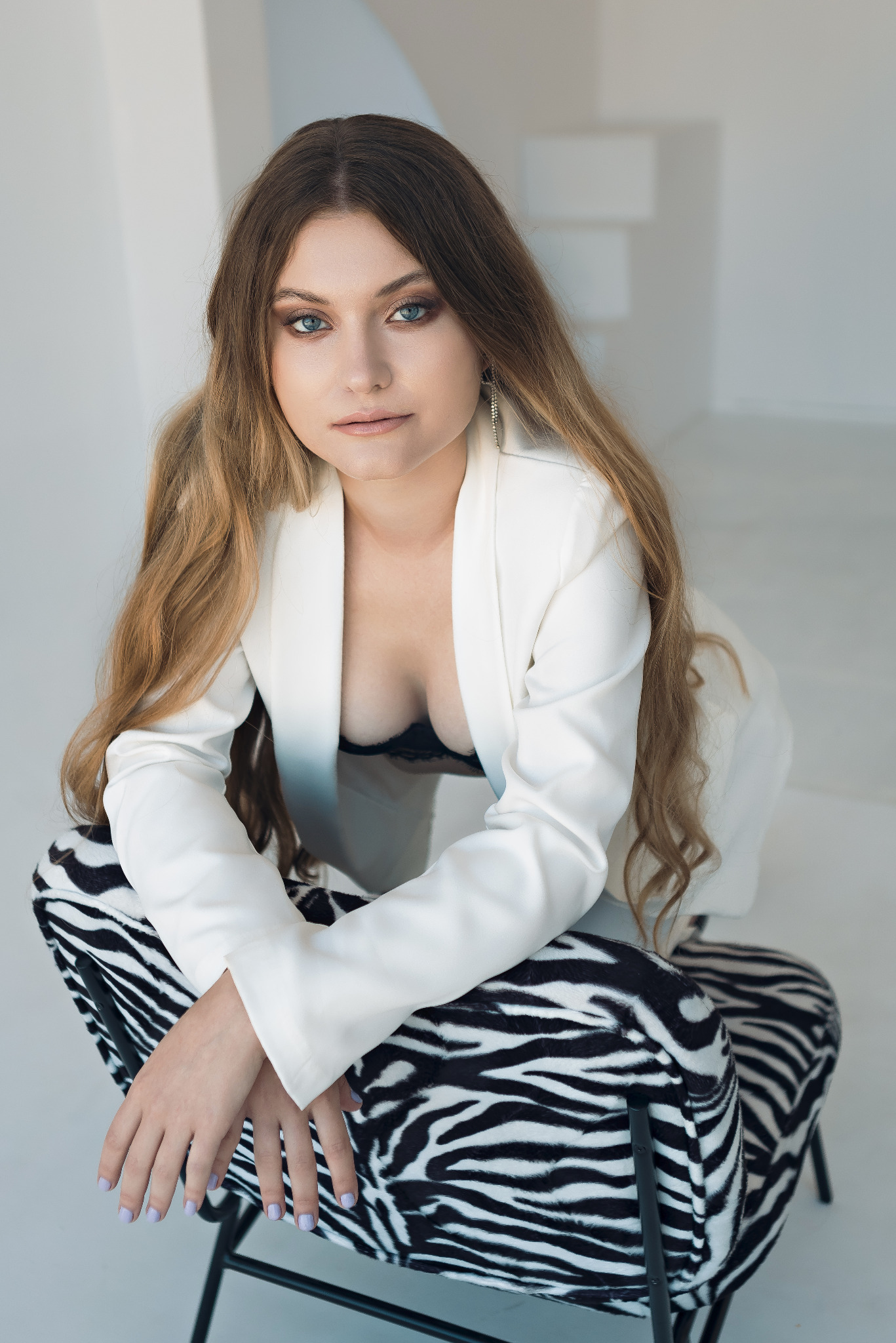 Portret kobiety w białym żakiecie, siedzącej na krześle w zebrę, z długimi blond włosami i delikatnym makijażem, patrzącej w obiektyw.