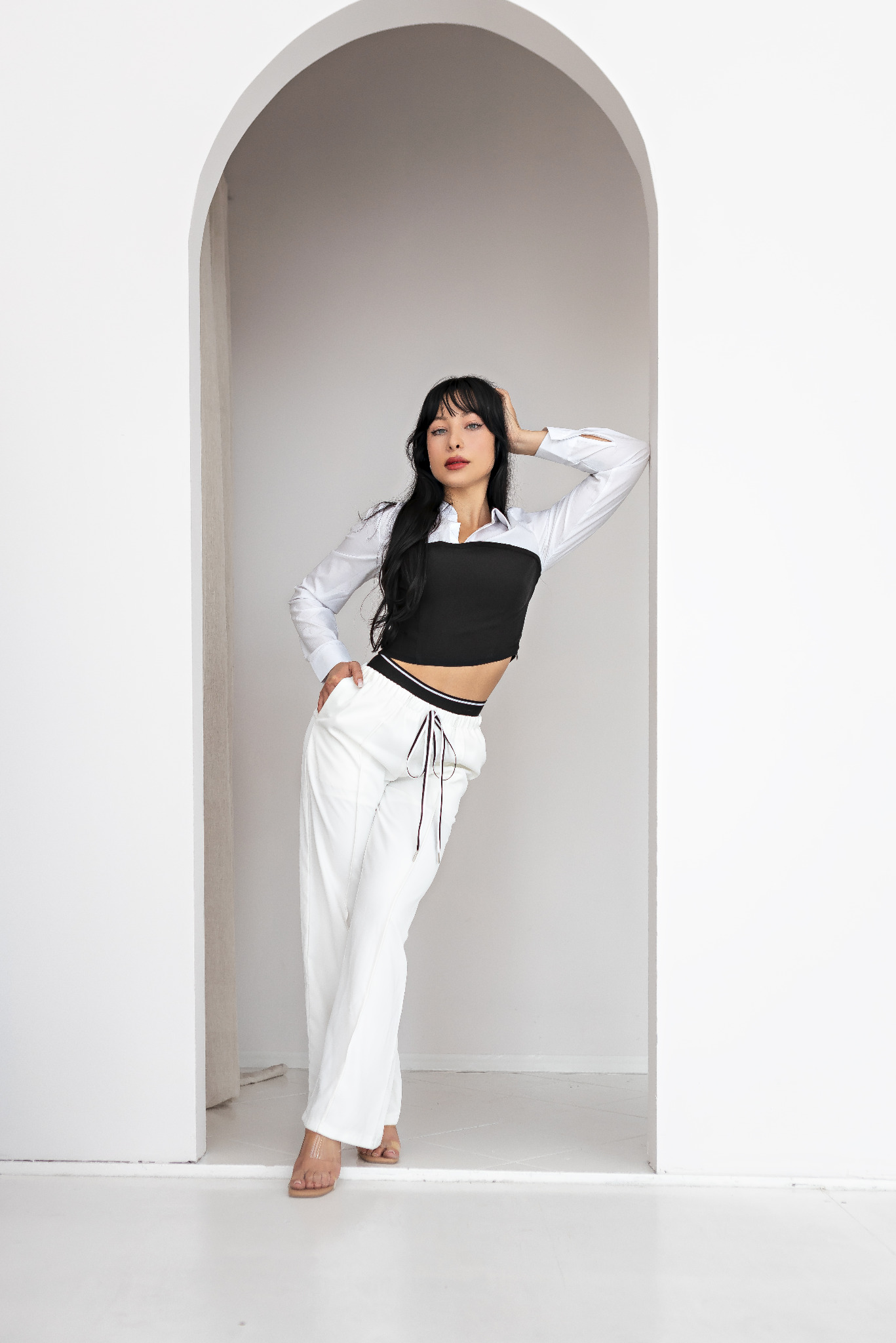 Kobieta w białych spodniach i czarnym gorsecie pozuje w białym, minimalistycznym wnętrzu z łukowym przejściem.