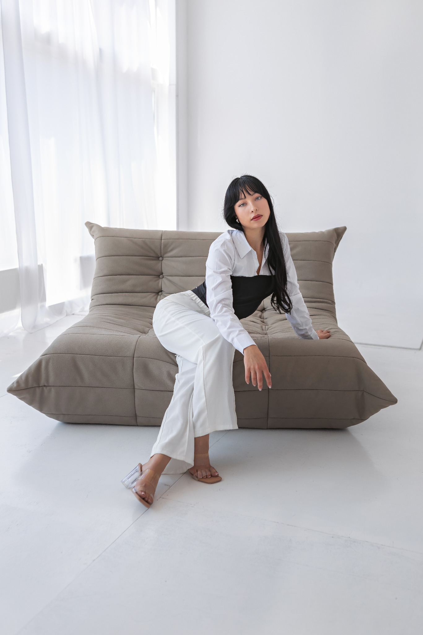 Kobieta z czarnymi włosami w białych spodniach i białej koszuli siedzi na beżowej, modułowej sofie na tle białej ściany i okna z białymi zasłonami. Jej dłoń spoczywa na sofie.