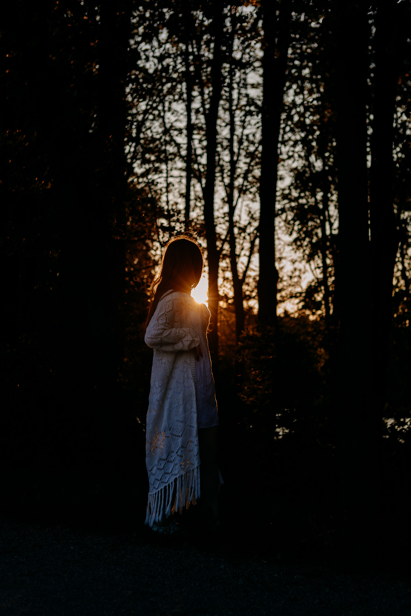 Sylwetka dziewczynki w lesie, podświetlona zachodzącym słońcem. Dziewczynka ubrana w biały sweter z frędzlami, stoi tyłem do obiektywu, patrząc w dal.