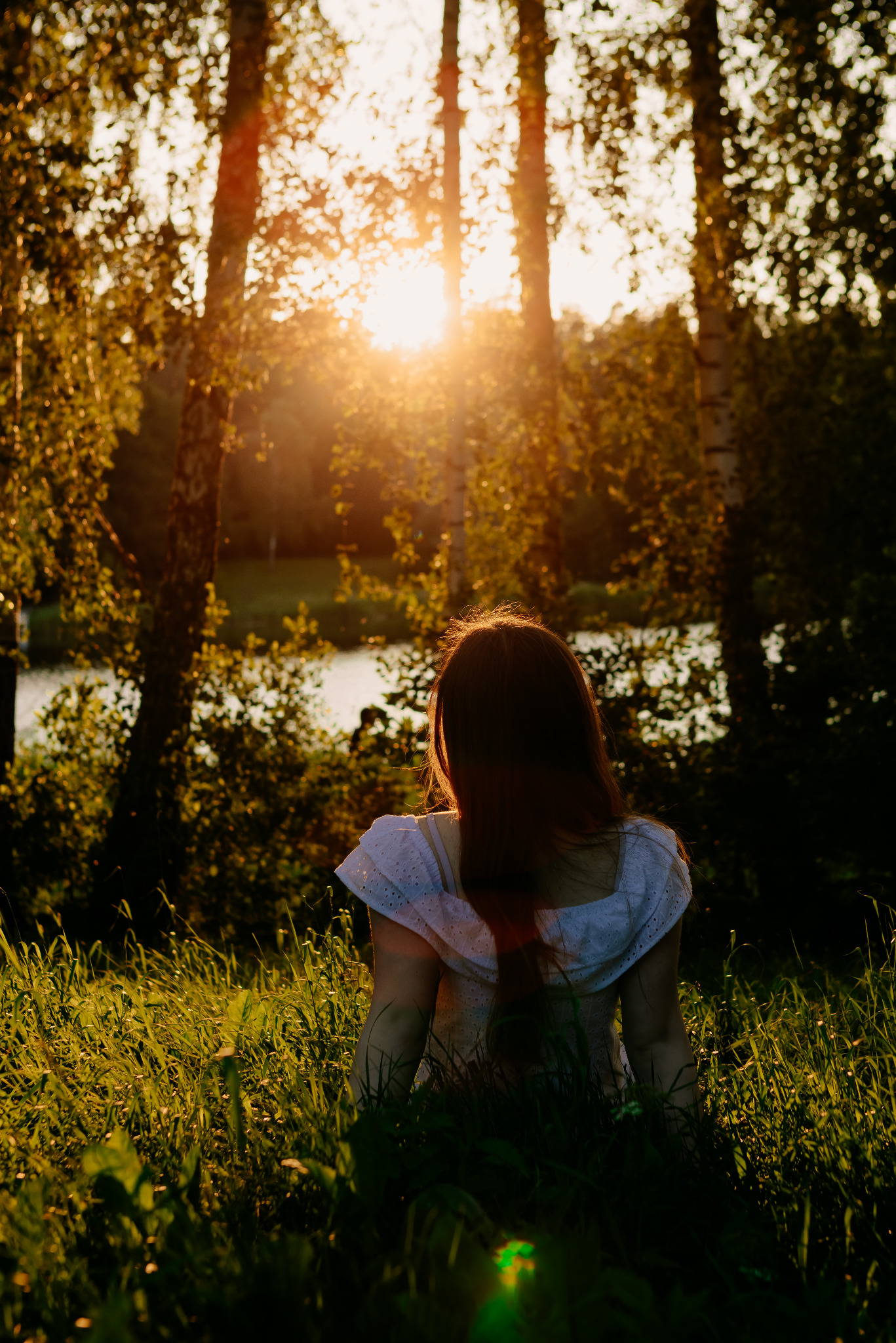 Kobieta w zwiewnej sukience siedzi w wysokiej trawie, plecami do widza, w tle drzewa i zachodzące słońce.