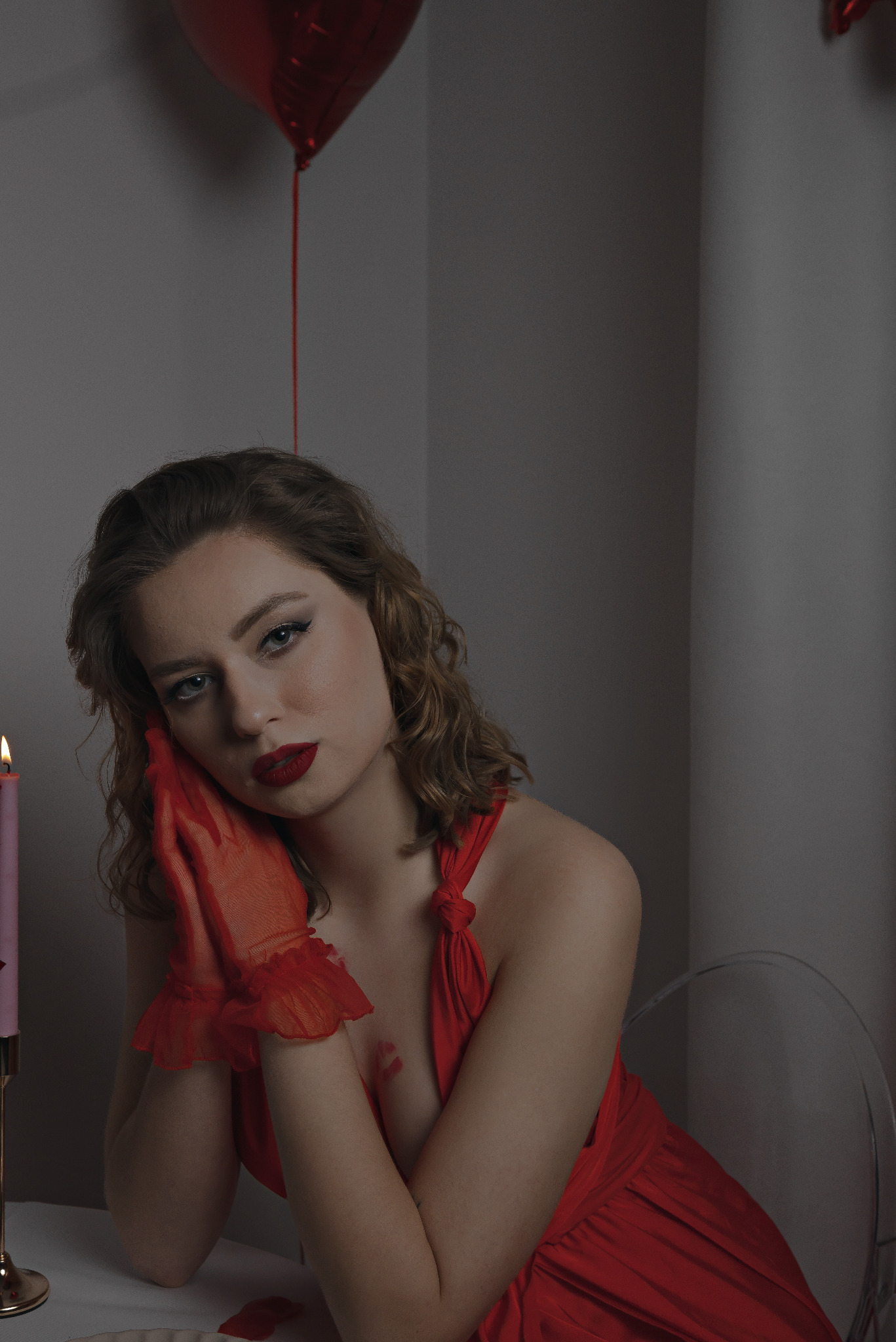 Portret kobiety w czerwonej sukience i rękawiczkach, z czerwonym balonem w tle, pozującej przy stole ze świecą. Romantyczna sesja zdjęciowa w studiu.