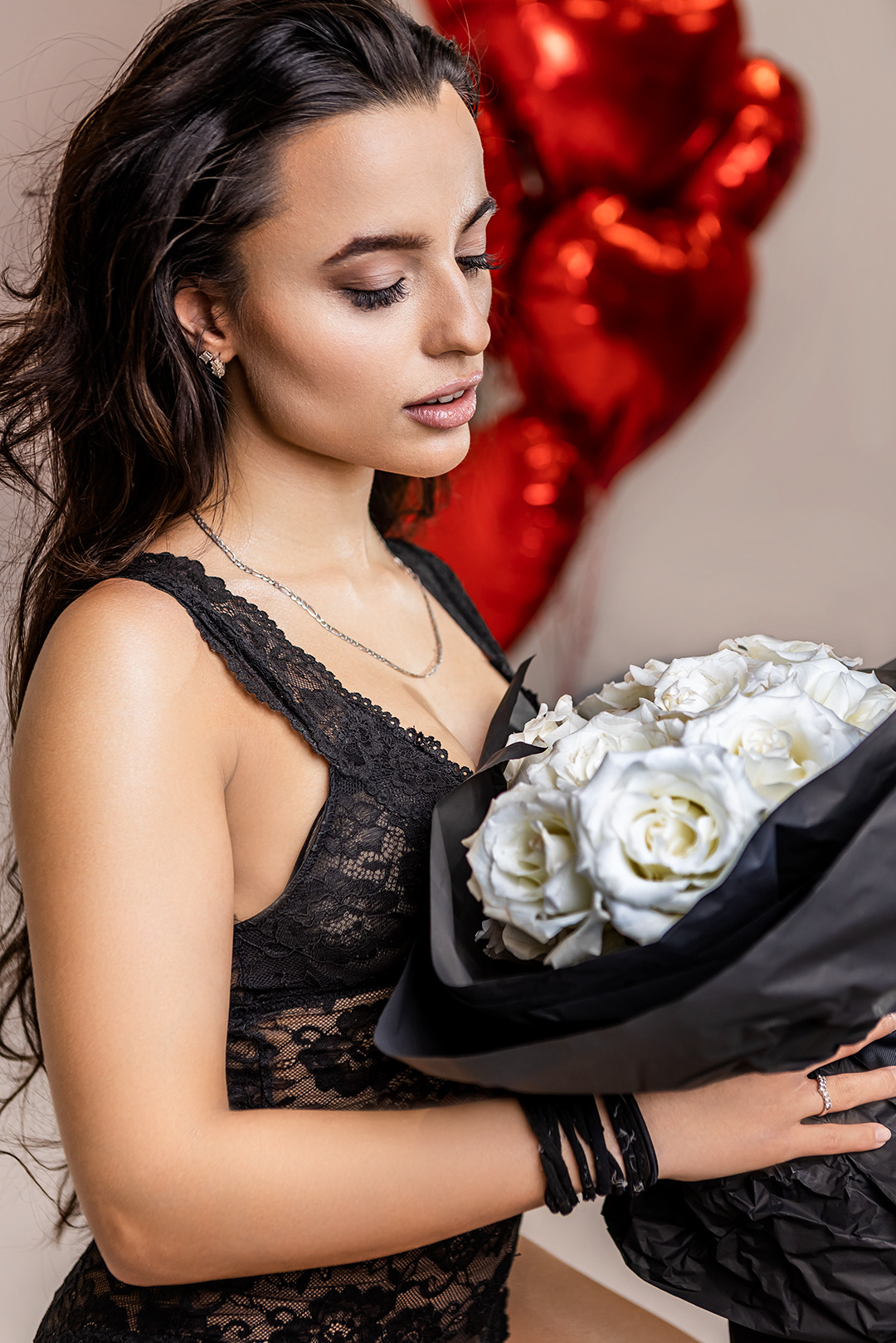 Zamyślona brunetka w czarnej bieliźnie trzyma bukiet białych róż na tle czerwonych balonów w kształcie serc. Romantyczna atmosfera, delikatne oświetlenie.