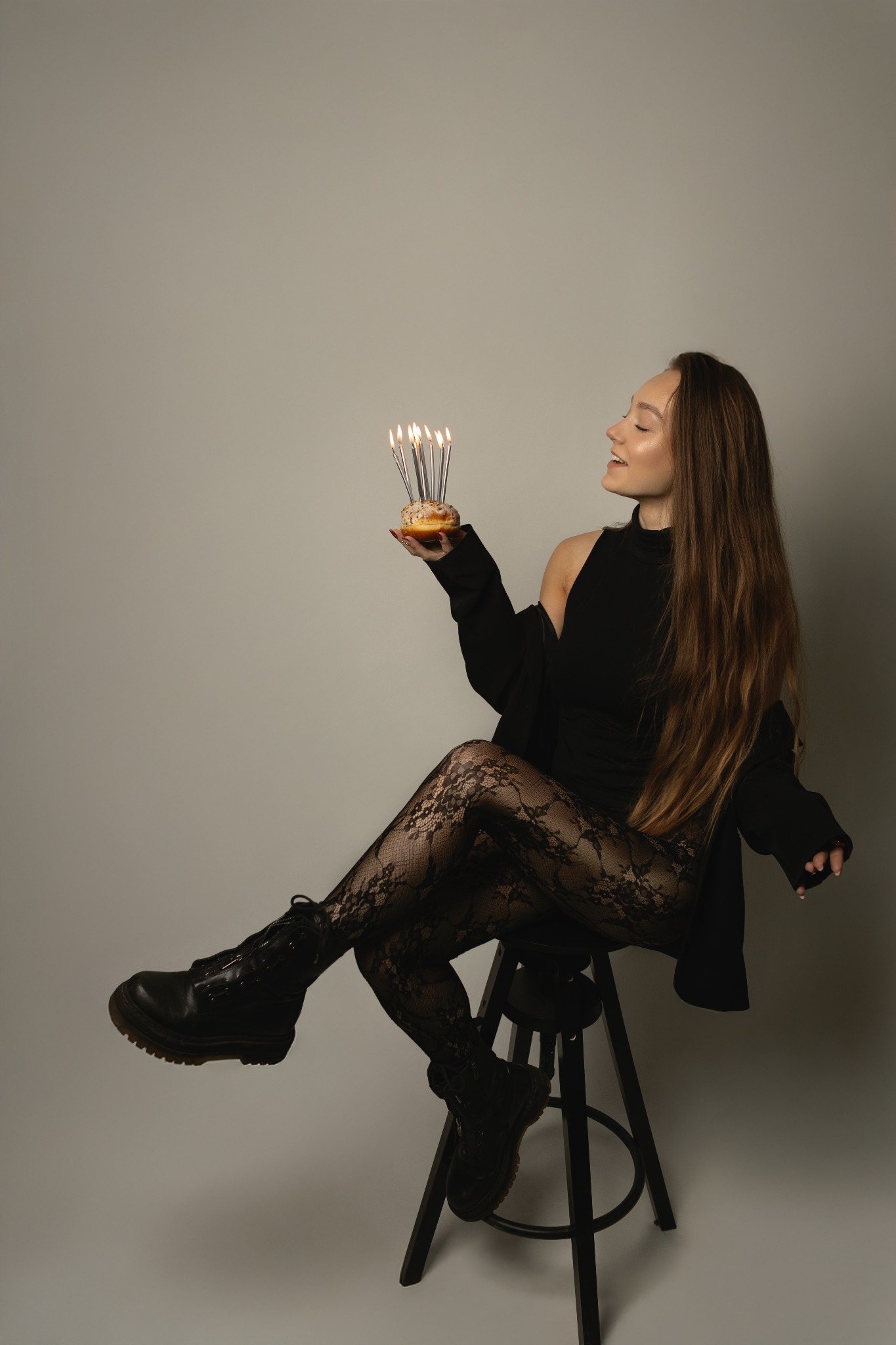 Kobieta w czarnym stroju siedzi na stołku, trzymając pączka ze świeczkami. Uśmiechnięta, z długimi włosami, w rajstopach w kwiaty i butach na platformie. Neutralne tło.