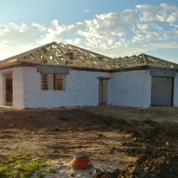 Surowy stan domu jednorodzinnego z białych bloków, w trakcie budowy dachu z drewnianą konstrukcją, na tle błękitnego nieba z chmurami.