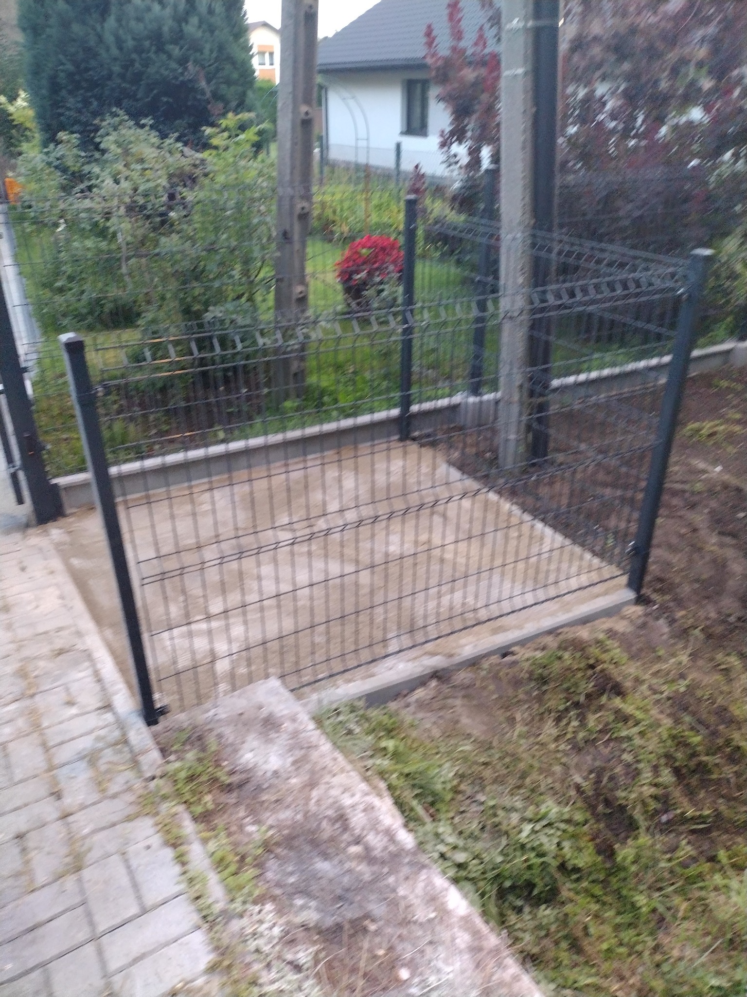 Nowo postawione, metalowe ogrodzenie panelowe na betonowej podmurówce, widok z góry na fragment posesji z kostki brukowej i zarośniętego trawnika.