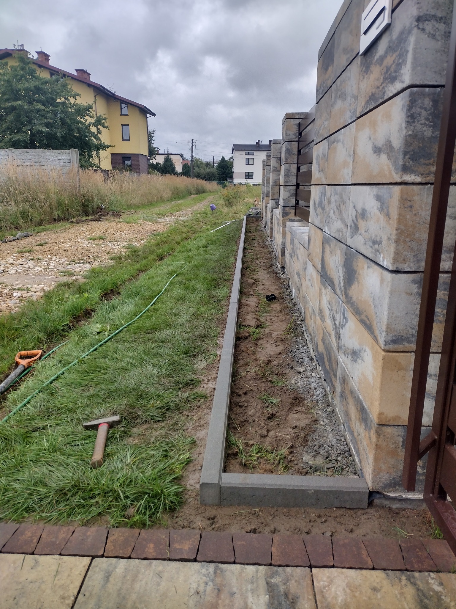 Wykopany rów obłożony obrzeżami betonowymi, przygotowany pod montaż ogrodzenia z betonowych bloczków, widoczny młotek i szpadel na trawie, domy w tle.