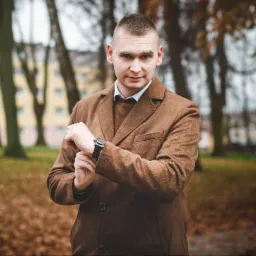 Mężczyzna w brązowym płaszczu, stojący na tle parku w Lidzbarku Warmińskim, sprawdza godzinę na czarnym zegarku, spoglądając w kierunku obiektywu.