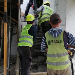 Eagle - Wylewanie betonowych schodów na budowie przez grupę robotników w kaskach i kamizelkach odblaskowych z logo firmy.