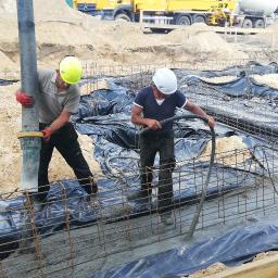 Eagle - Wylewanie betonu do fundamentów zbrojonych stalą, zabezpieczonych folią, widoczni trzej pracownicy w kaskach i rękawicach, w tle betonowóz i hałdy piasku.