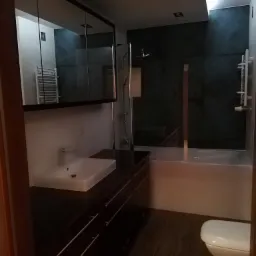 Nowoczesna łazienka z ciemnymi płytkami imitującymi kamień, podwieszaną szafką z umywalką, lustrem w zabudowie i toaletą w rogu pomieszczenia.