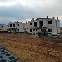 Eagle - Surowe, dwukondygnacyjne domy w zabudowie szeregowej w trakcie budowy, wykonane z szarego betonu komórkowego, z widocznymi blokami betonu na dachach i paletach, oraz pracownikami na dachu jednego...