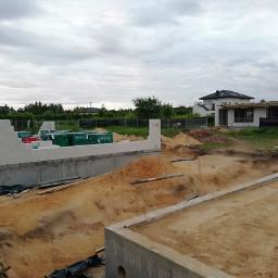 Eagle - Wczesny etap budowy domu: fundamenty z betonu i pierwsze warstwy bloczków z betonu komórkowego, widoczny wykop pod kolejne fundamenty, w tle dom w stanie surowym otwartym i sąsiednie domy.