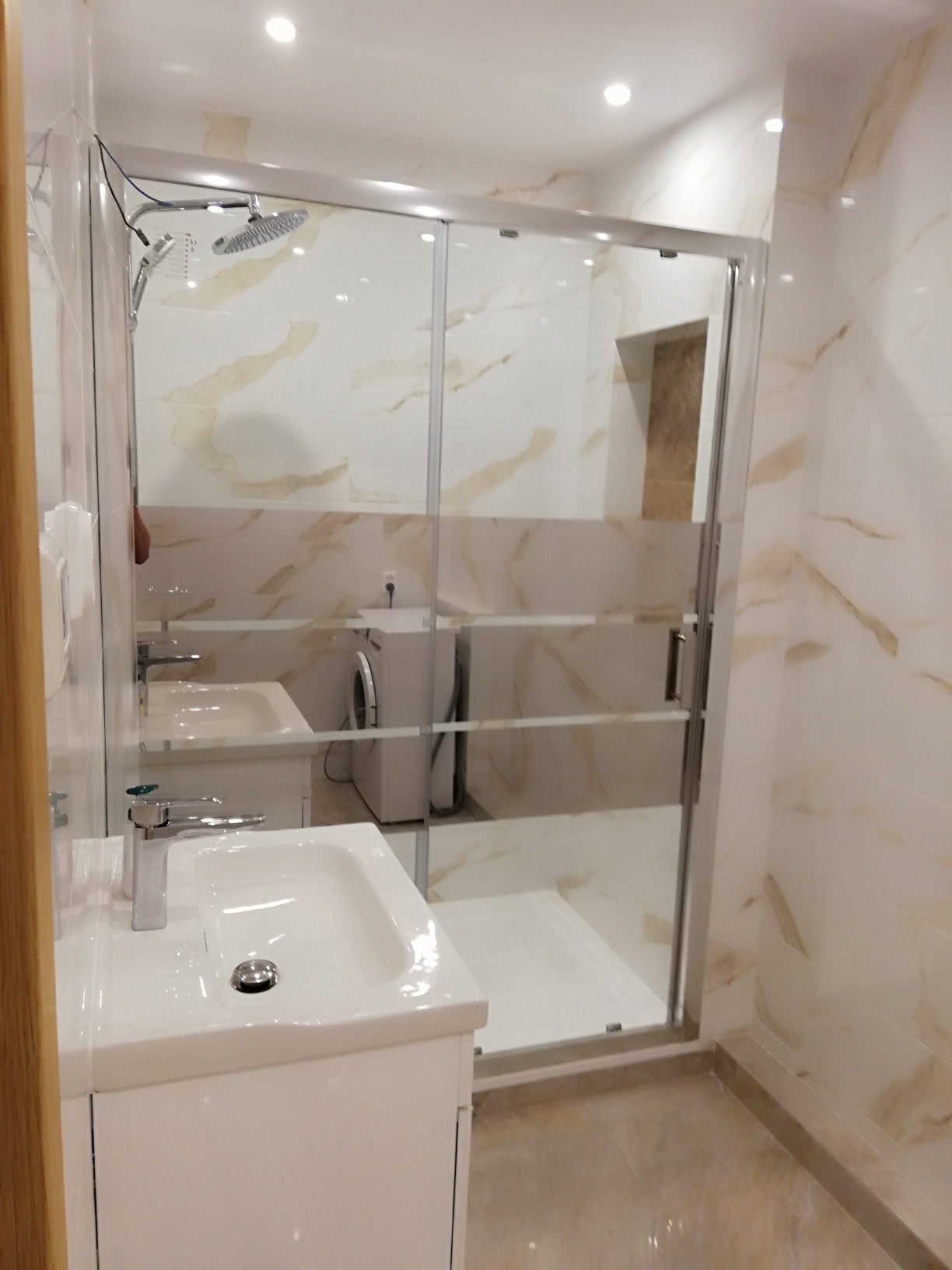Nowoczesna łazienka z kabiną prysznicową typu walk-in, umywalką na szafce i pralką w tle, wykończona płytkami imitującymi marmur w odcieniach beżu i bieli.