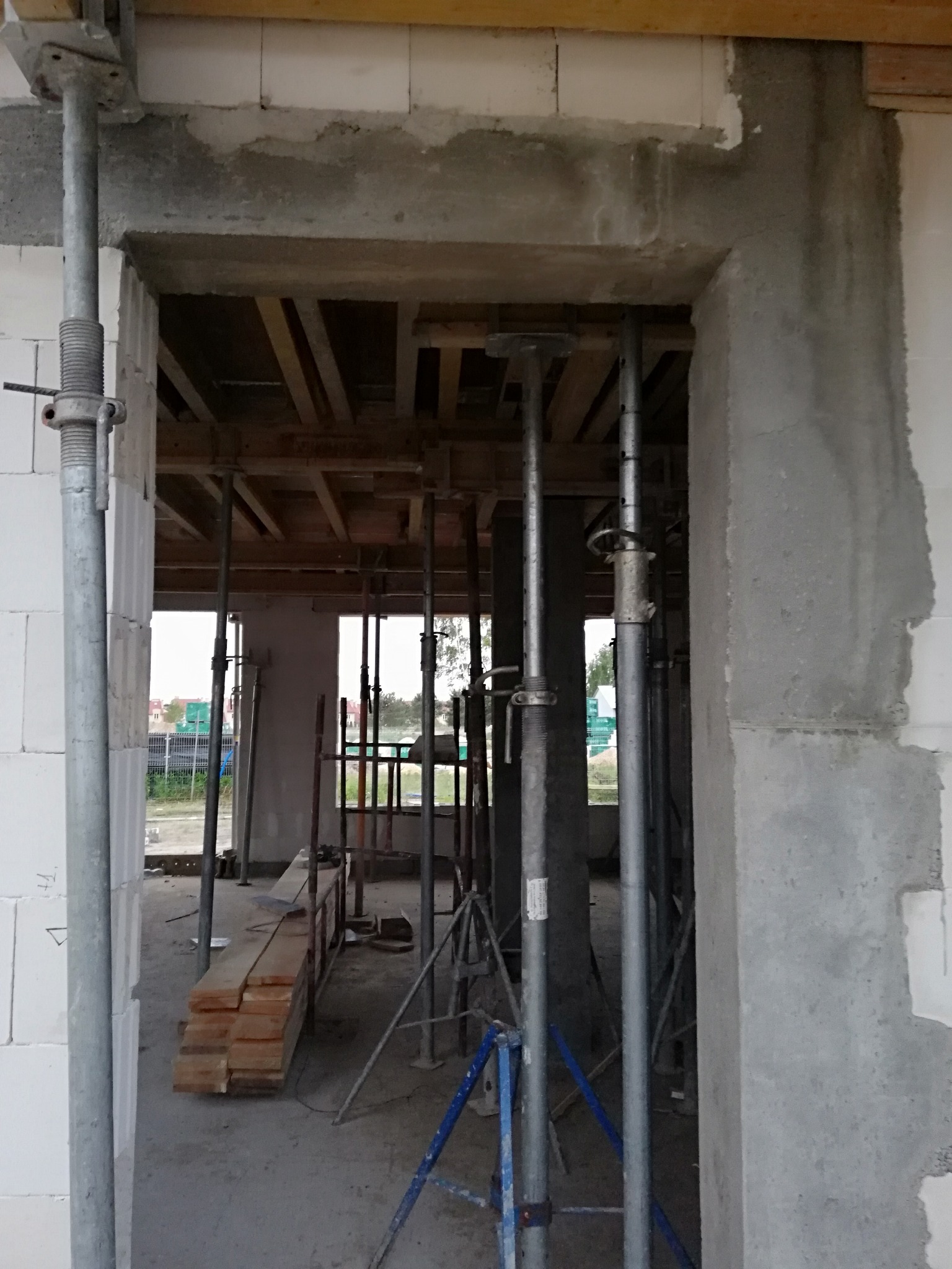 Surowy stan budowy domu: widok przez otwór drzwiowy na drewniany strop podparty metalowymi stemplami, ściany z białych bloczków i szarych betonowych słupów.