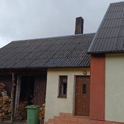 Pokrycia Dachowe Paweł Poniatowski - Dom z szarym dachem z eternitu, widoczne ślady mchu. Stos drewna pod zadaszeniem. Pochmurne niebo, lekki deszcz. Ujęcie z poziomu gruntu.