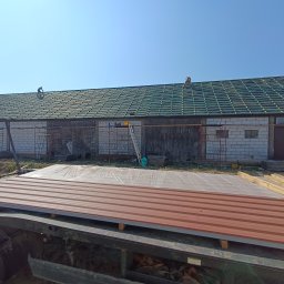 Pokrycia Dachowe Paweł Poniatowski - Montaż zielonego dachu z blachy trapezowej na budynku gospodarczym. Na dachu pracują dwie osoby, a na przyczepie leżą arkusze blachy w kolorze ceglastym.