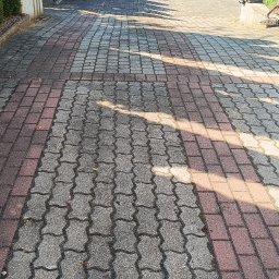 Pokrycia Dachowe Paweł Poniatowski - Nawierzchnia z szarych falistych i czerwonych prostokątnych elementów chodnikowych, z widocznymi zabrudzeniami i mchem na szarych. W tle różowy budynek z czerwonymi drzwiami garażowymi.