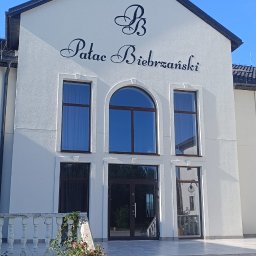 Pokrycia Dachowe Paweł Poniatowski - Biała fasada Pałacu Biebrzańskiego z ciemnymi oknami i dachem, widoczne schody, rabata kwiatowa oraz fragment urządzenia do czyszczenia powierzchni na jasnej kostce brukowej.