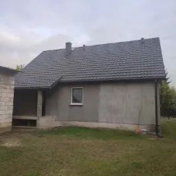 Dom w stanie surowym z nowym, grafitowym dachem z blachodachówki, widoczne obróbki blacharskie i komin, na trawniku stoi wiadro z farbą i grabie.