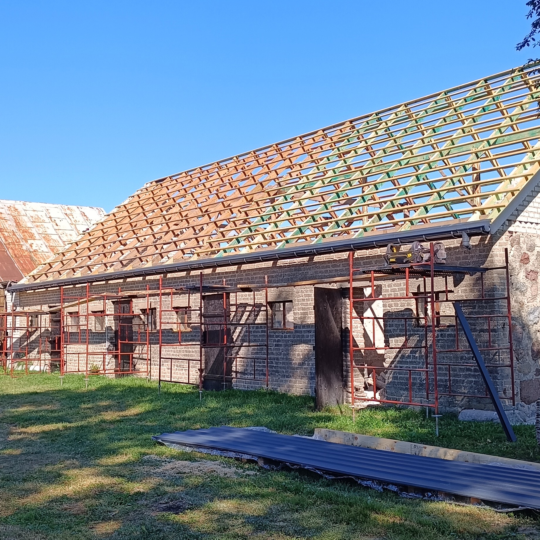 Budynek z cegły w trakcie wymiany dachu. Widoczne rusztowania, konstrukcja dachu z drewnianych łat i kontrłat, oraz przygotowane arkusze blachy na trawniku.