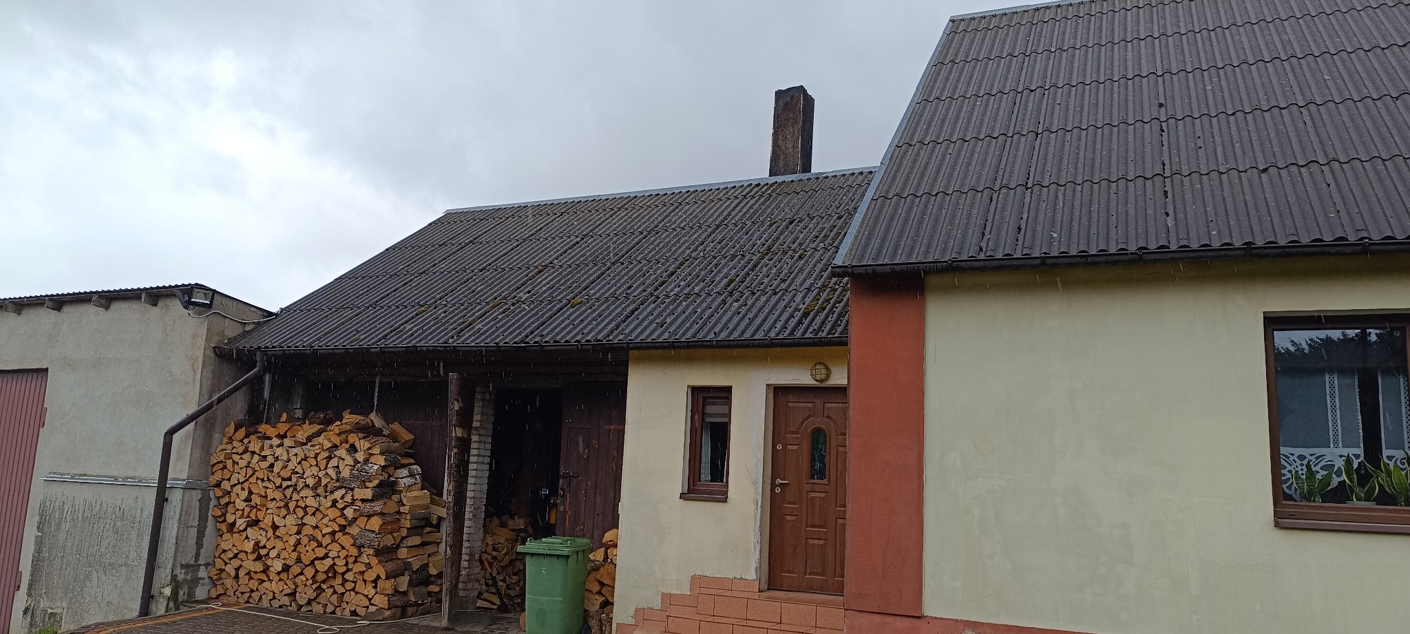 Dom z szarym dachem z eternitu, widoczne ślady mchu. Stos drewna pod zadaszeniem. Pochmurne niebo, lekki deszcz. Ujęcie z poziomu gruntu.