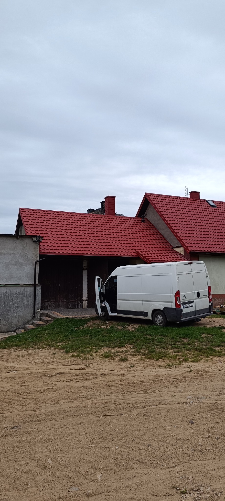 Dom z nowym, czerwonym dachem z blachodachówki trapezowej, zaparkowany biały bus z otwartymi drzwiami. Widok z poziomu gruntu, pochmurne niebo.