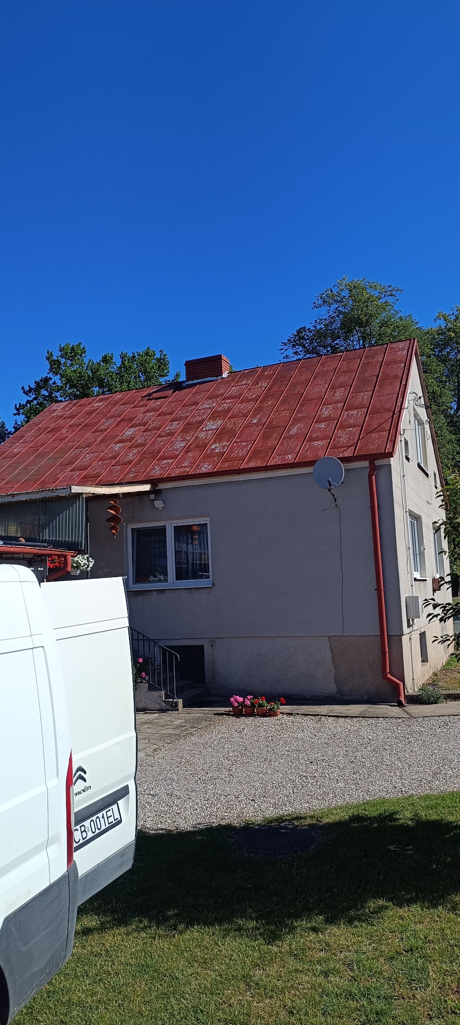 Dom z czerwonym dachem z blachy trapezowej, widoczny z boku, z zaparkowanym białym busem. Na dachu antena satelitarna.