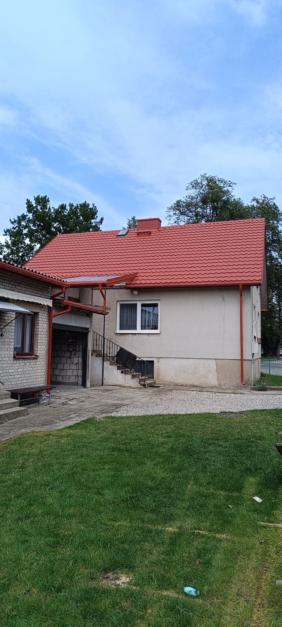 Dom z nowym, czerwonym dachem z blachy trapezowej i rynnami w kolorze dachu. Widoczny fragment elewacji i otoczenie budynku: trawnik, drzewa.