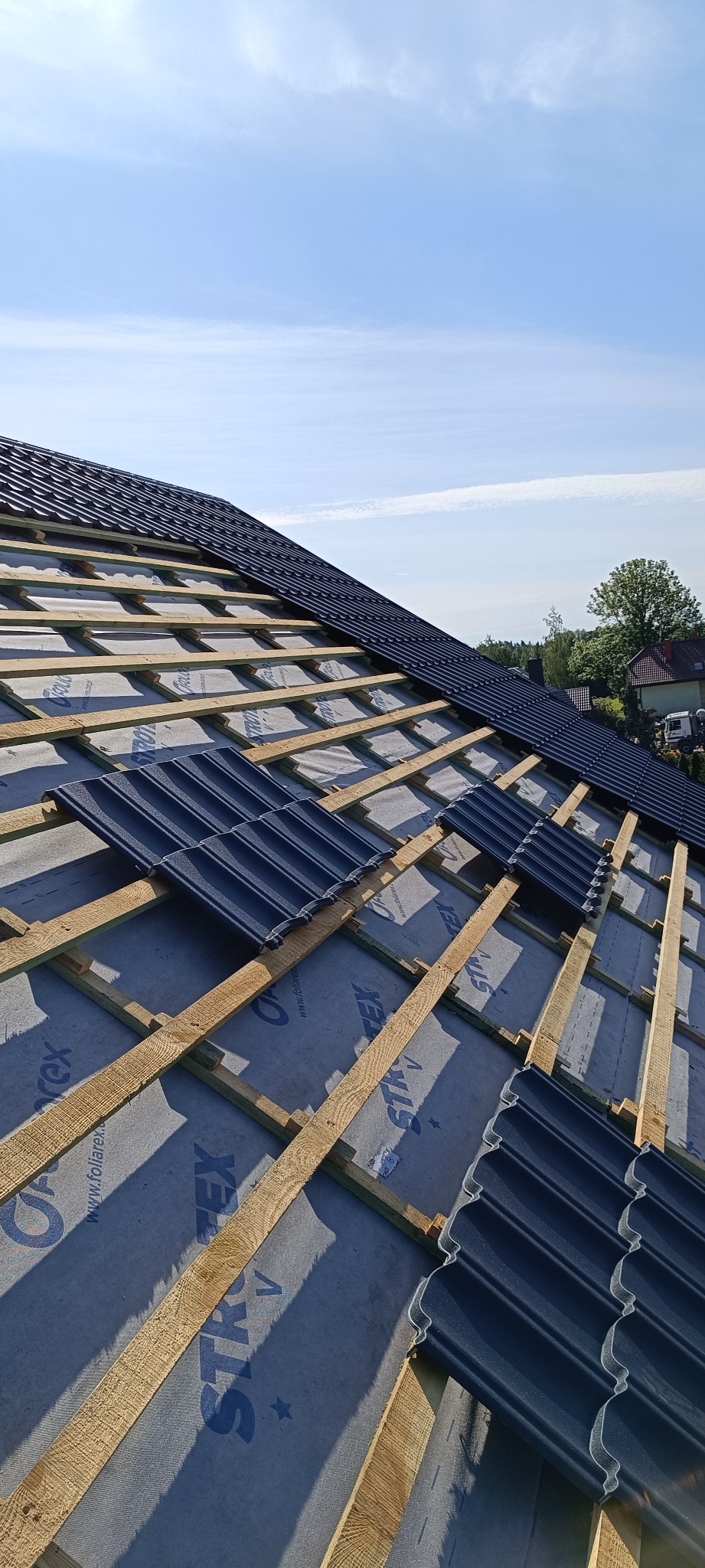 Montaż ciemnej dachówki na łatach i membranie dachowej. Widok z góry, częściowo pokryty dach w słoneczny dzień. Detal konstrukcji dachu.