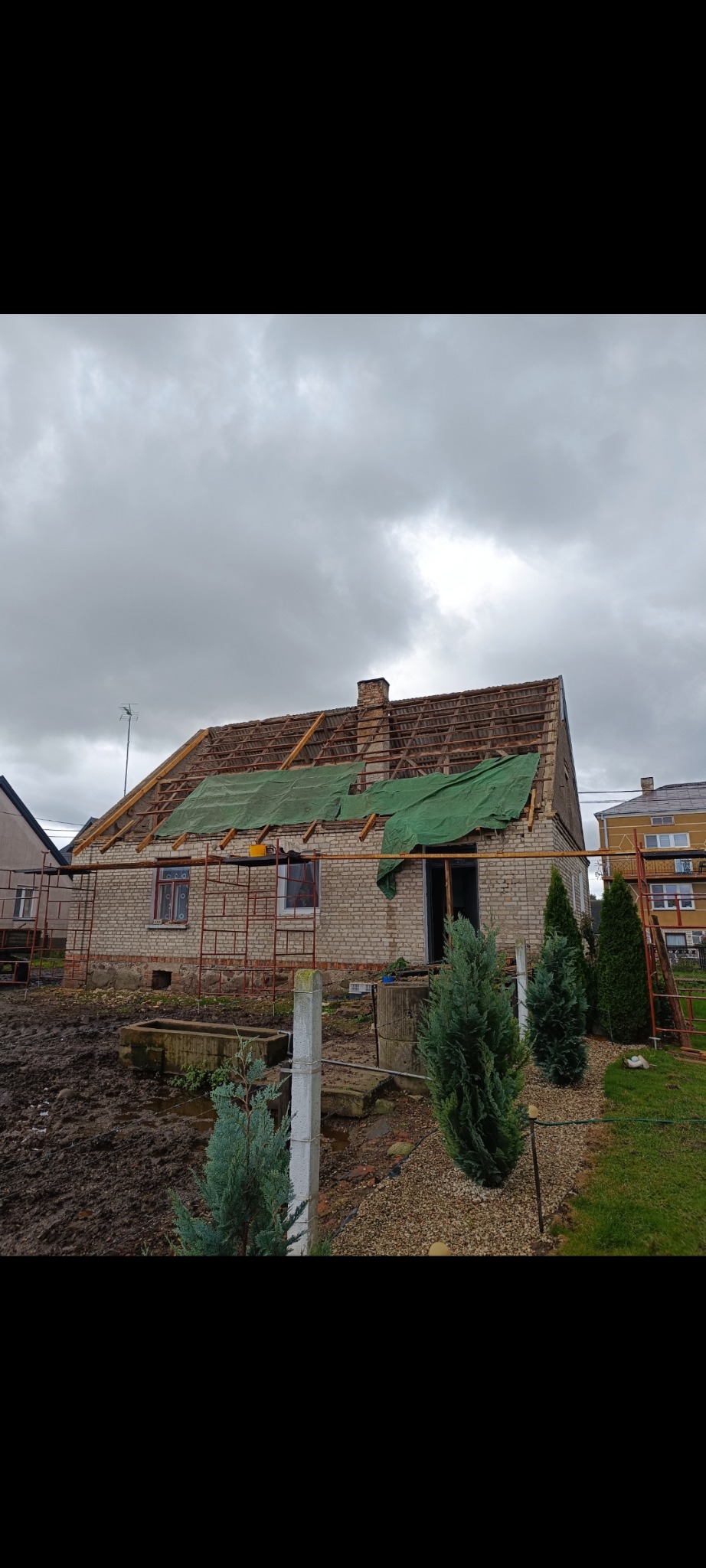Ceglany dom w trakcie wymiany dachu, widoczne rusztowania i odsłonięta konstrukcja. Część dachu przykryta zieloną plandeką. Pochmurne niebo w tle.