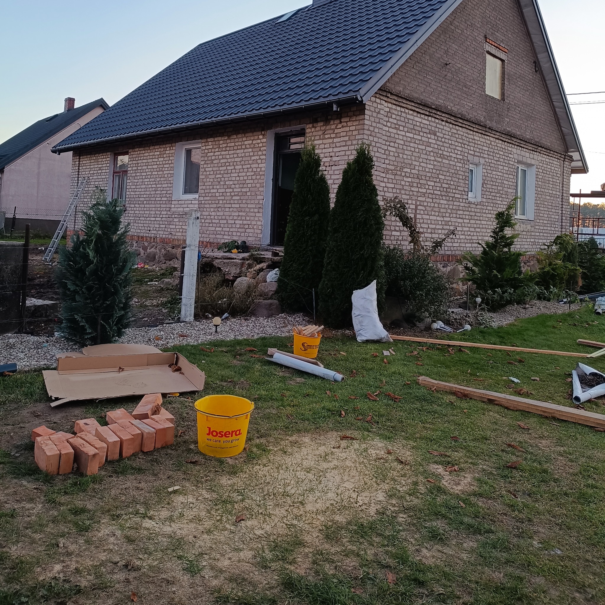 Dom z ciemną dachówką w trakcie prac wykończeniowych ogrodu. Widoczne materiały budowlane, rośliny i narzędzia. Ujęcie z poziomu trawnika.