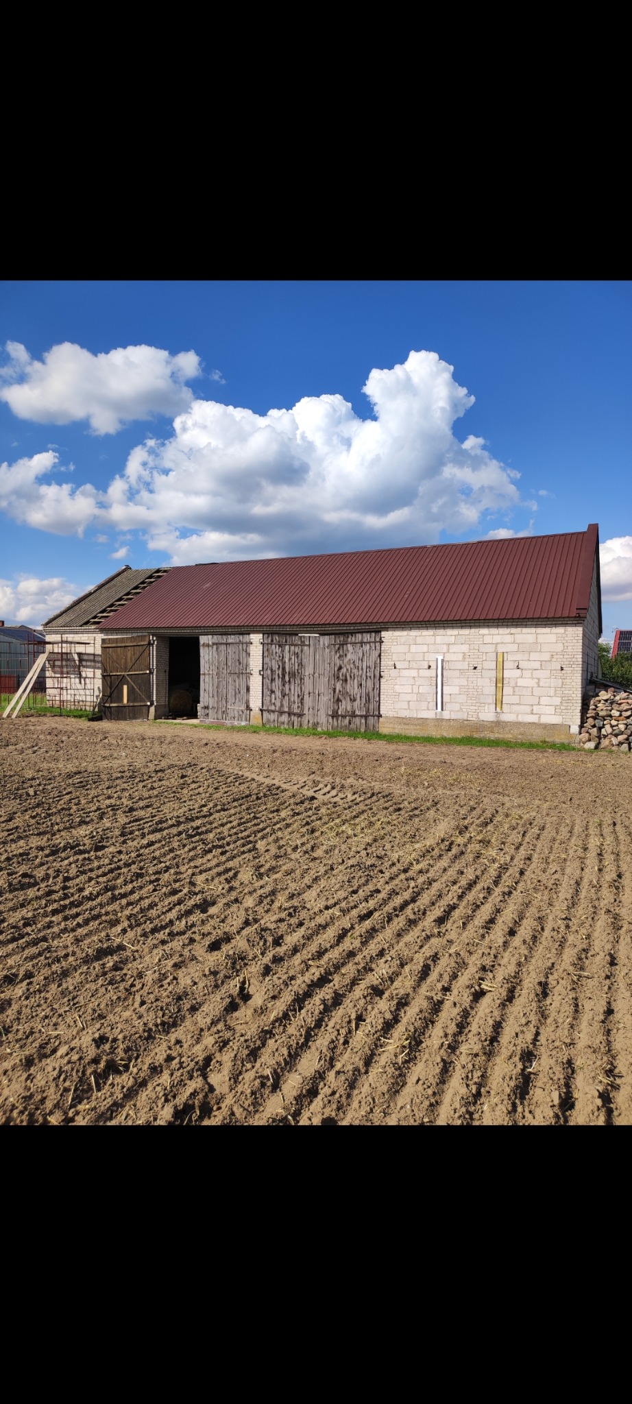 Budynek gospodarczy z czerwonym dachem, częściowo uszkodzonym, na tle błękitnego nieba z chmurami. Z przodu pole z bruzdami.