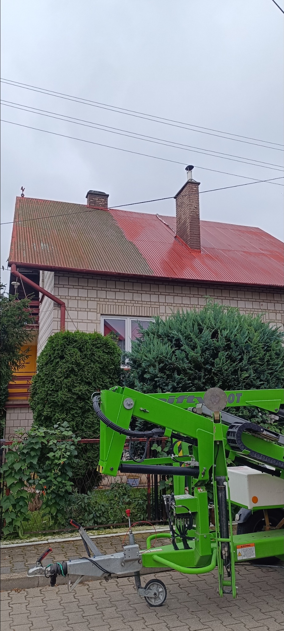 Dom z częściowo odnowionym, czerwonym dachem z blachy trapezowej. Widoczny fragment zielonego podnośnika koszowego. Część dachu pokryta mchem. Pochmurne niebo.