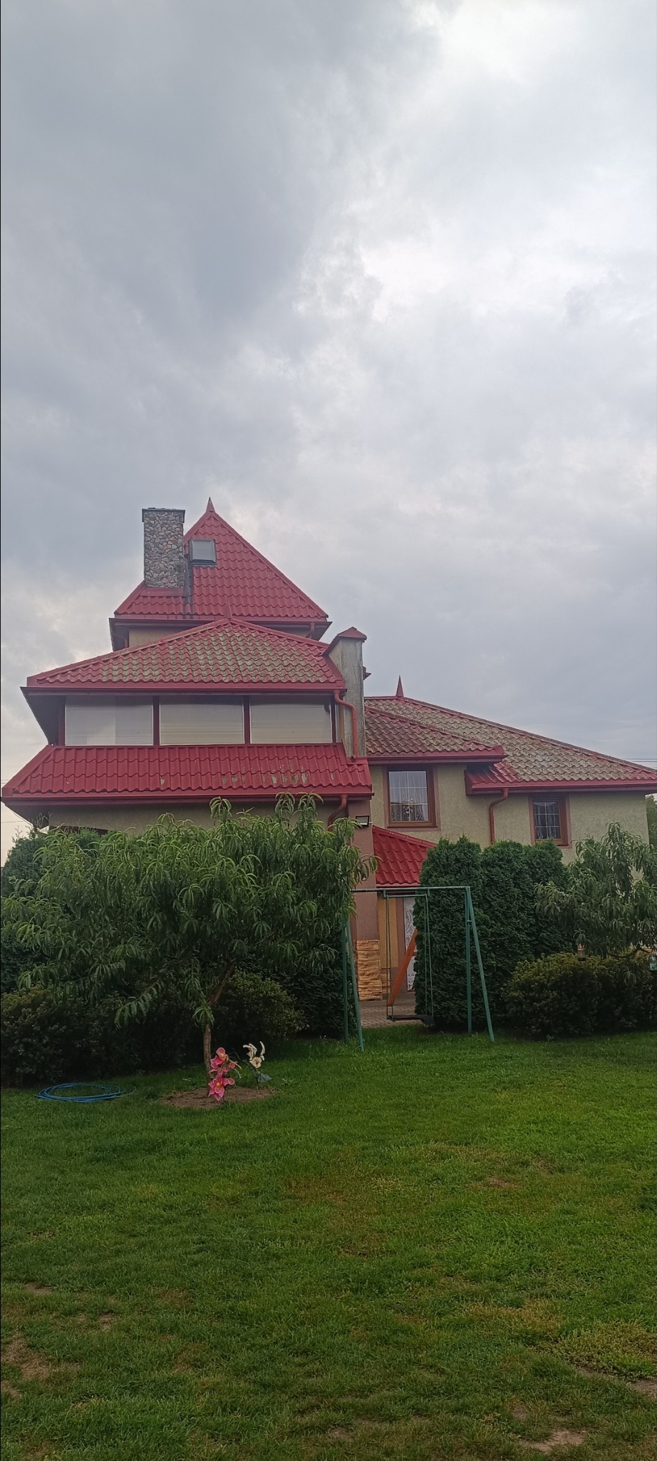 Dom z czerwonym dachem wielospadowym i kamiennym kominem, otoczony zielenią. Widok na elewację i fragment ogrodu z huśtawką. Pochmurne niebo w tle.