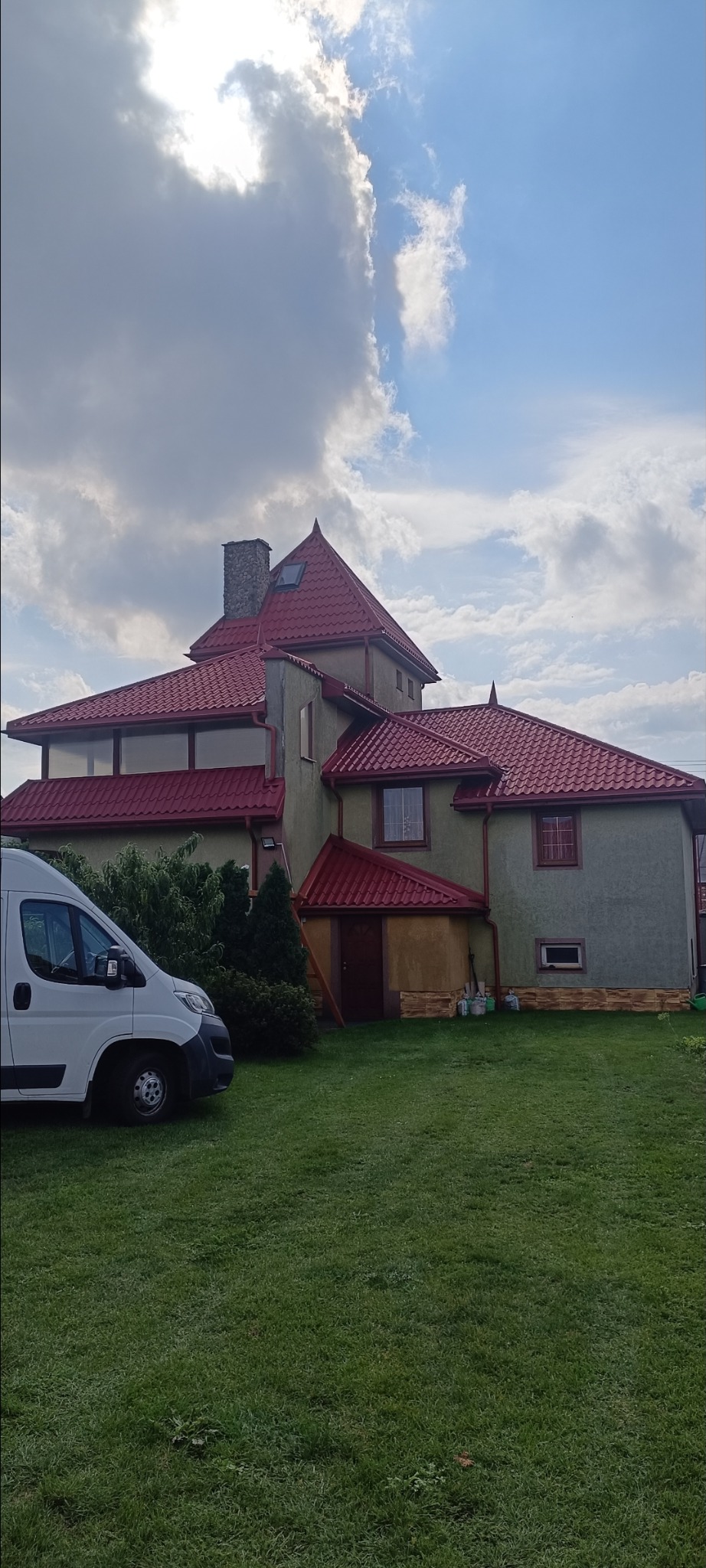 Dwupiętrowy dom z czerwonym dachem i szarymi ścianami, widziany z poziomu trawnika. Biały van zaparkowany obok. Częściowo pochmurne niebo w tle.