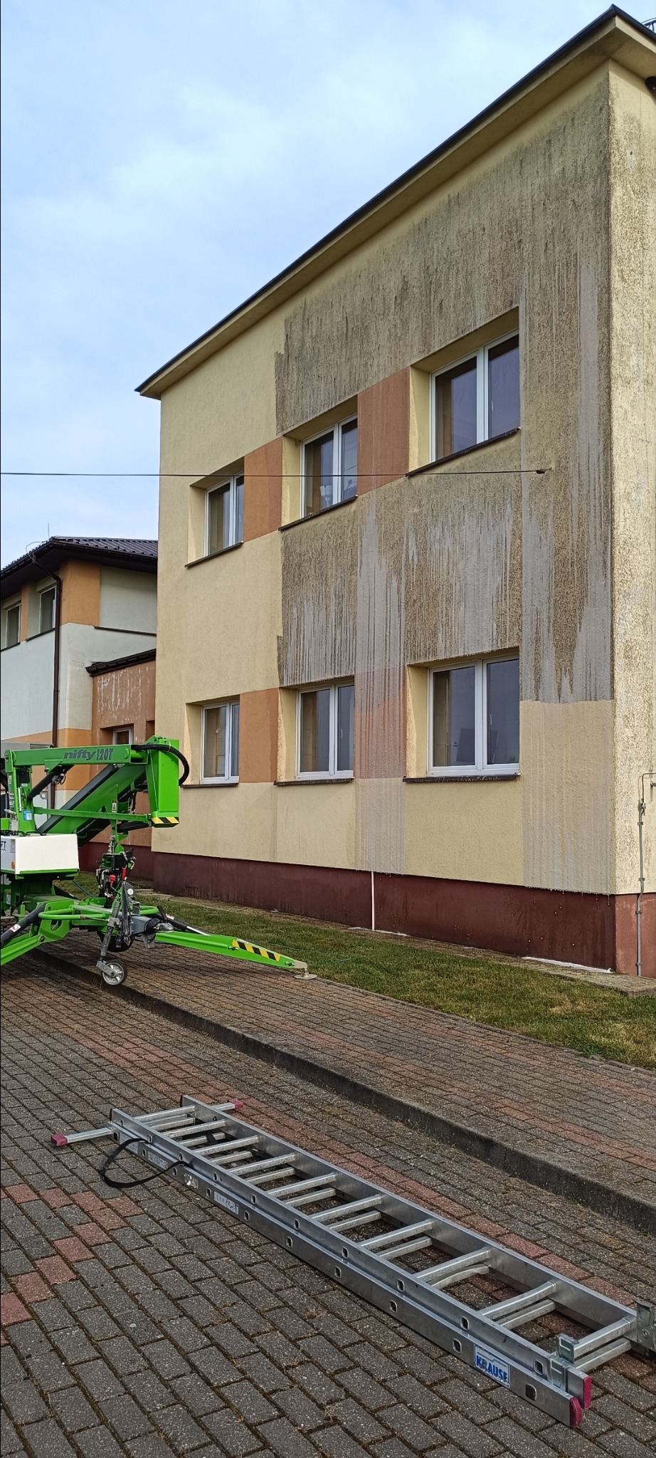 Fragment elewacji budynku z widocznymi śladami mycia, z użyciem podnośnika koszowego i drabiny. Częściowo umyta ściana ukazuje różnicę w kolorze.