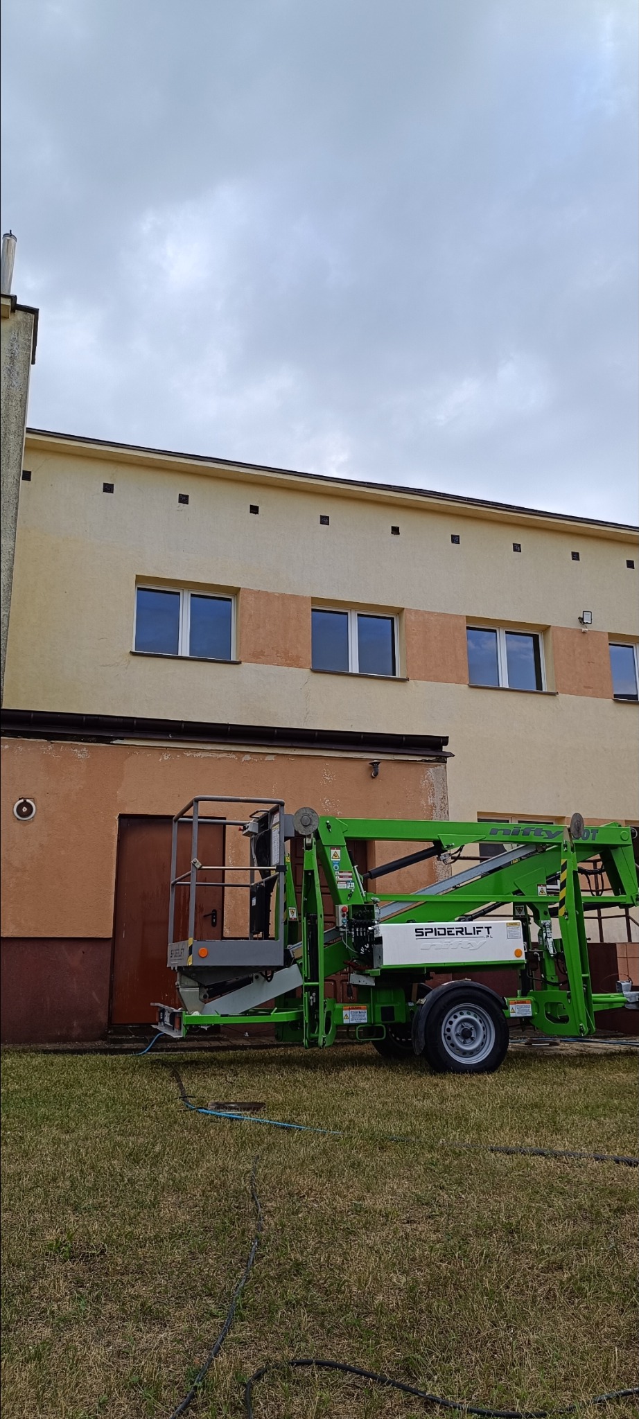 Zielony podnośnik koszowy Spiderlift na trawie przed budynkiem z beżową elewacją i brązowymi drzwiami garażowymi. Sprzęt gotowy do pracy.