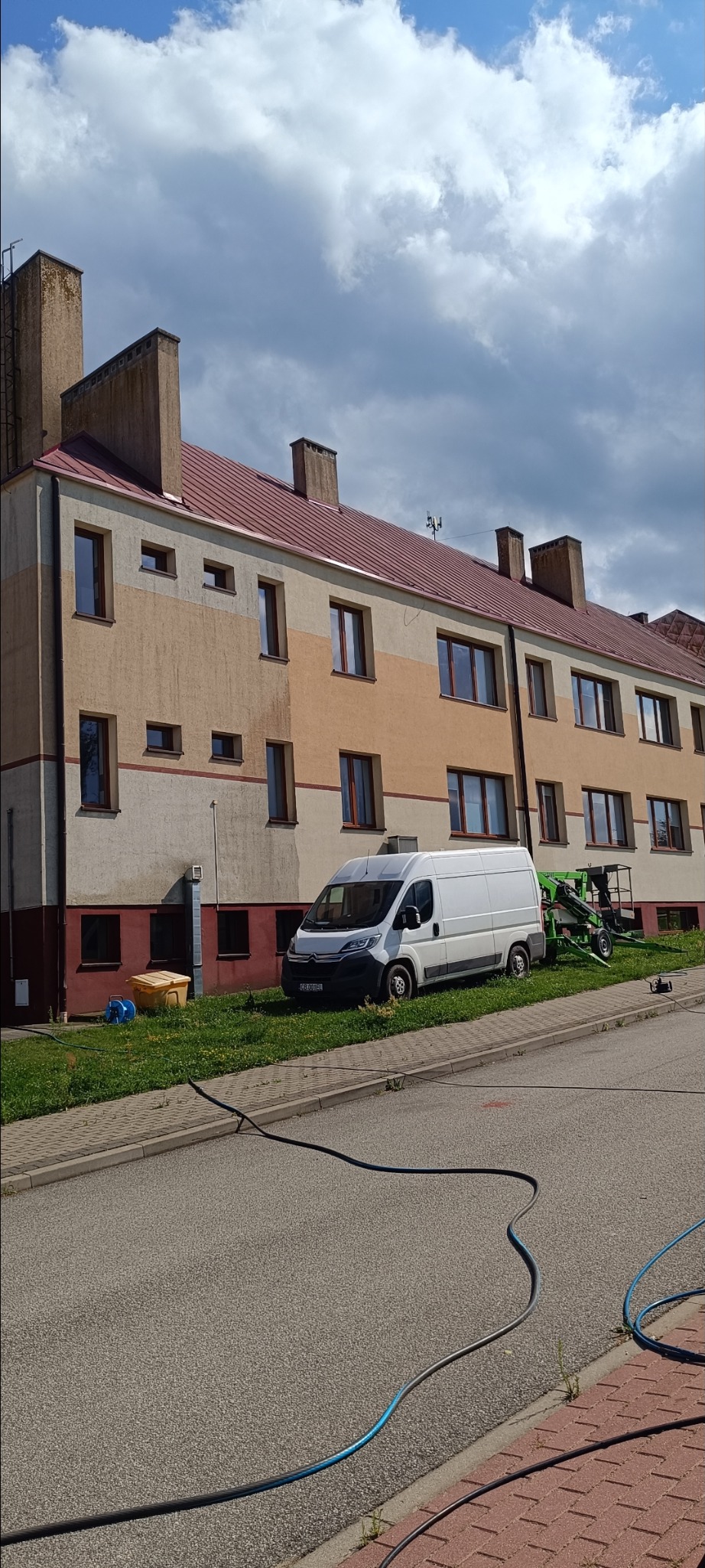Wielorodzinny budynek z nowym dachem, biały bus i podnośnik koszowy na trawniku. Węże na chodniku sugerują mycie elewacji. Słoneczny dzień, lekkie zachmurzenie.