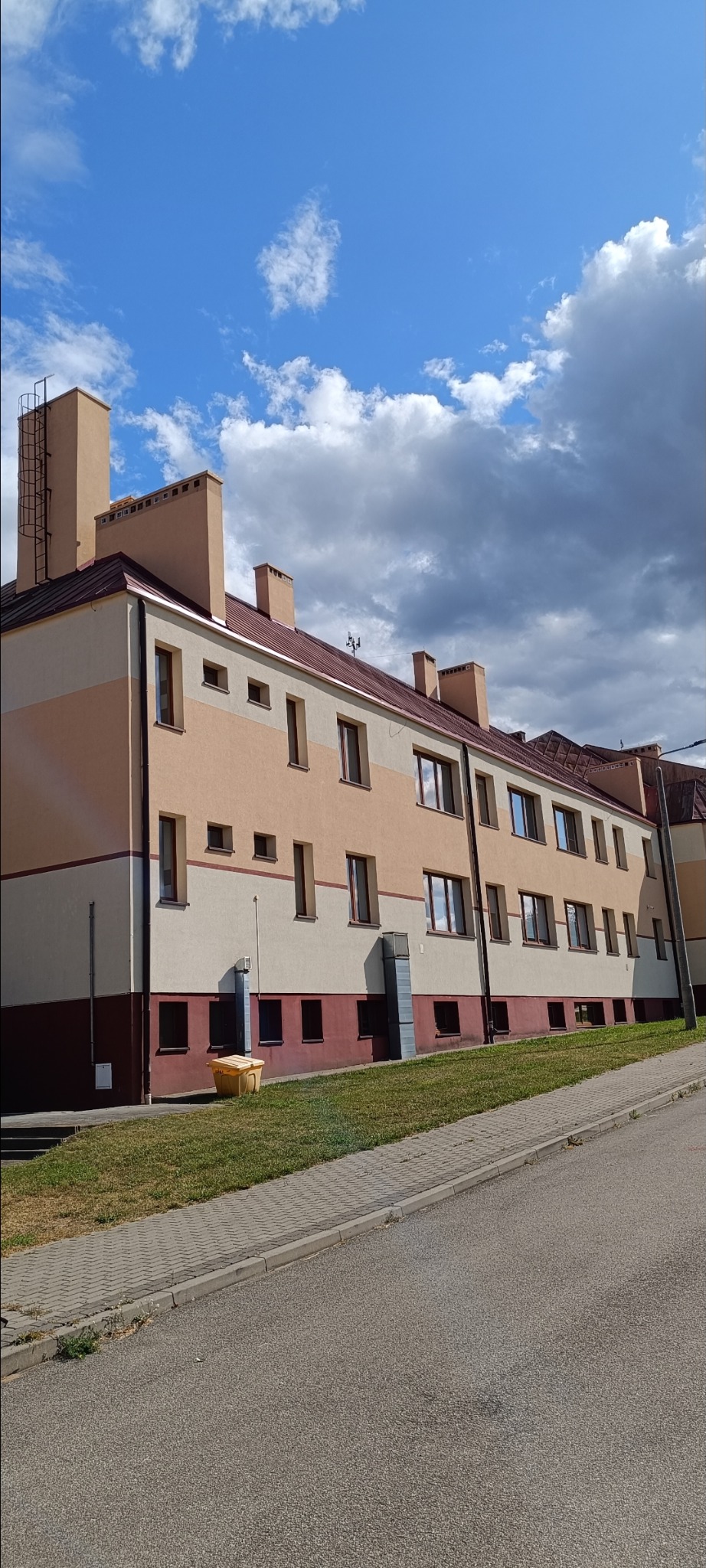 Wielopiętrowy budynek po renowacji elewacji, z nowym dachem i rynnami, na tle błękitnego nieba z chmurami. Widok z poziomu ulicy, z żółtym pojemnikiem na śmieci.