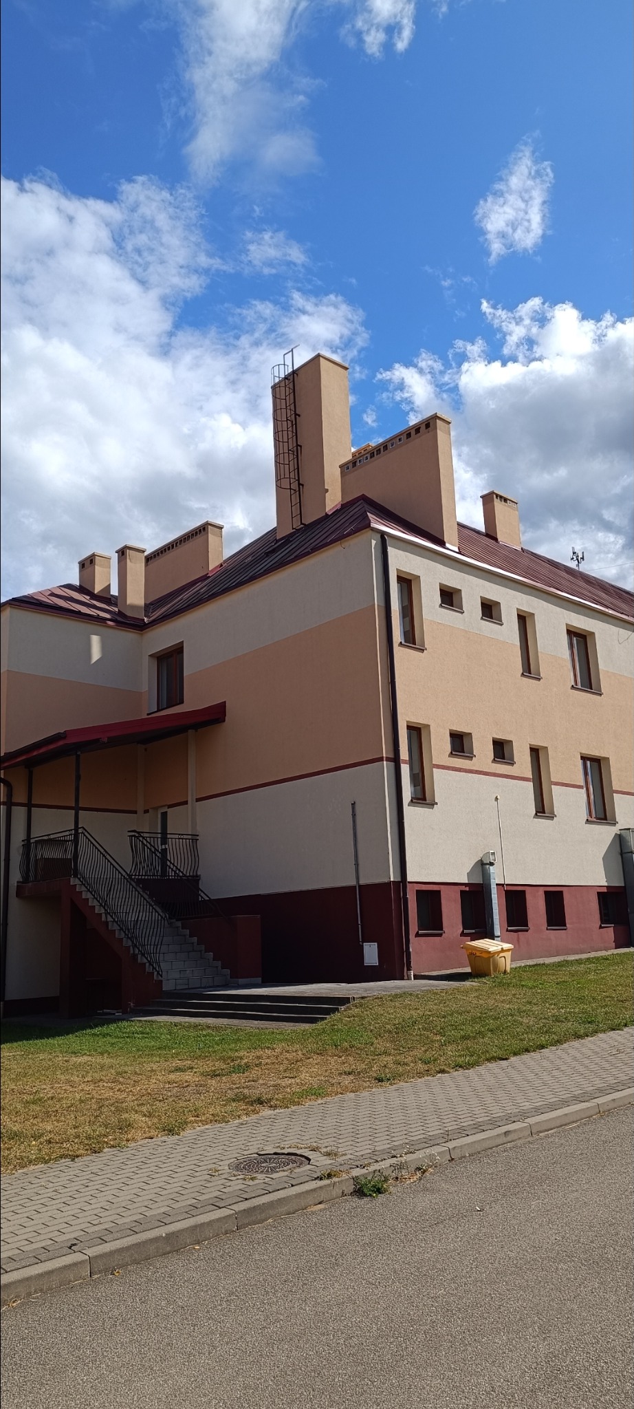 Dwupiętrowy budynek z beżową elewacją i brązowym dachem, widoczny z poziomu chodnika. Na dachu kominy z drabinką. Schody wejściowe z czarną balustradą.