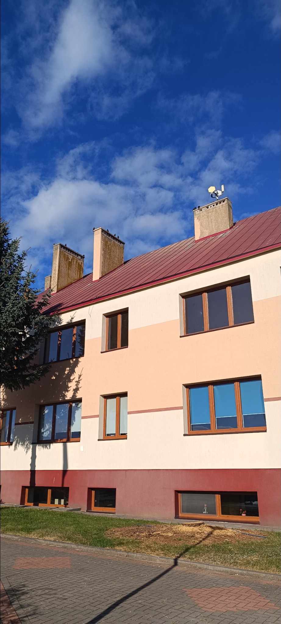 Wielopiętrowy budynek z czerwonym dachem i jasną elewacją w odcieniach beżu, widziany z dołu pod kątem. Na dachu kominy i antena. Błękitne niebo z chmurami w tle.