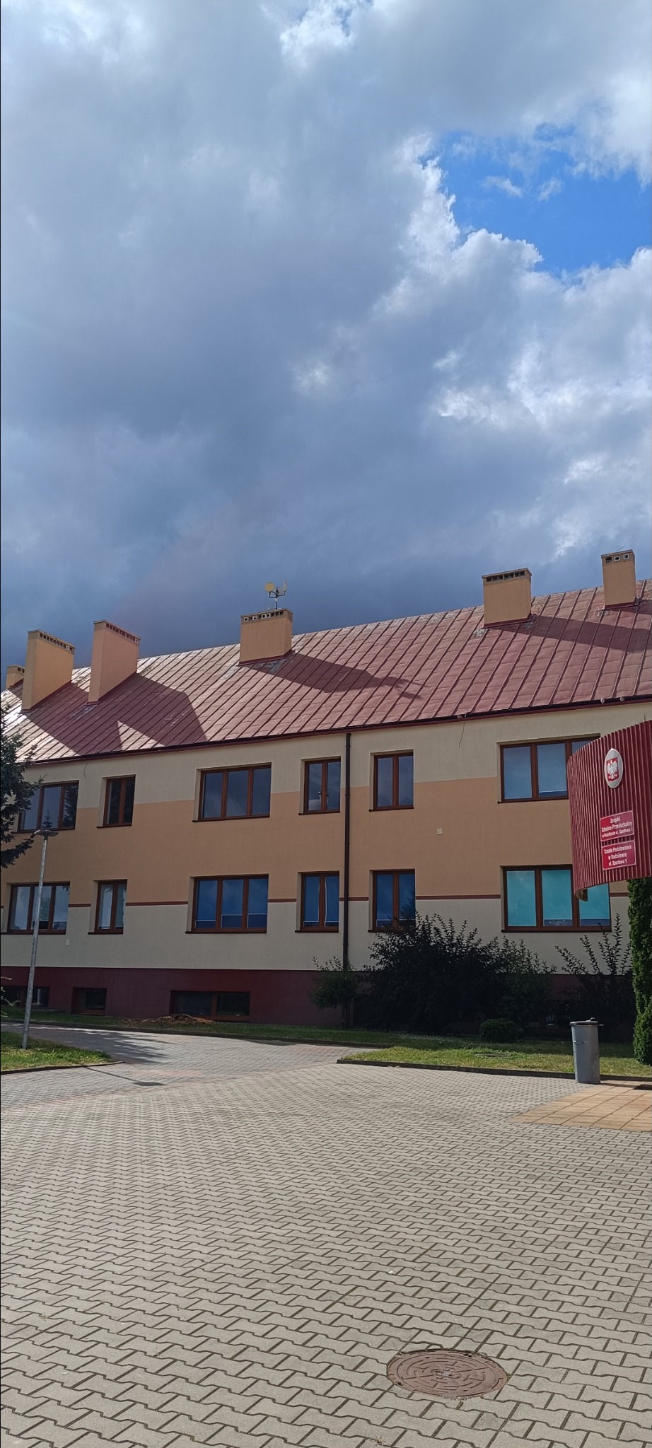 Budynek z czerwoną dachówką i beżową elewacją pod pochmurnym niebem. Widoczny fragment kostki brukowej i zieleni. Na dachu antena.