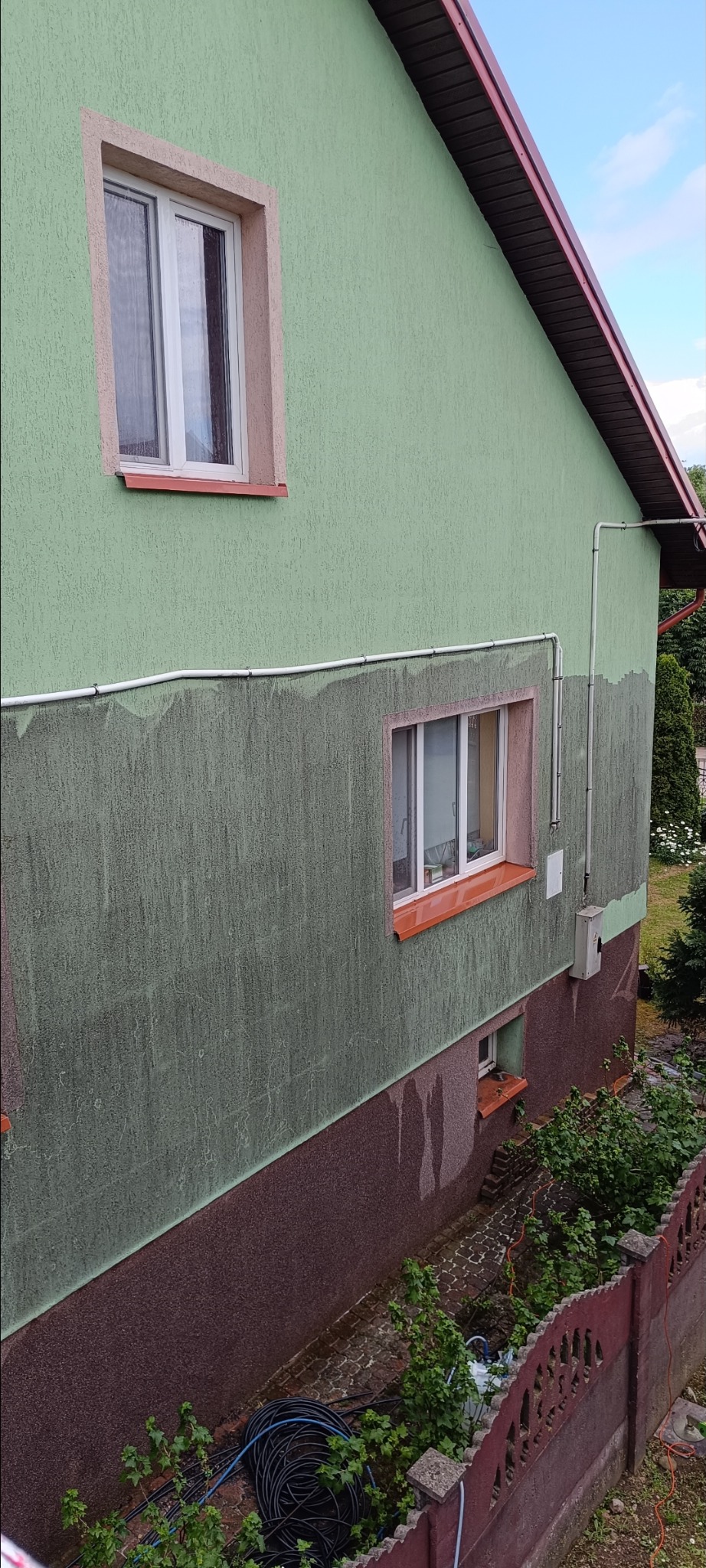 Fragment ściany budynku z widocznymi śladami po myciu elewacji. Górna część ściany jest czysta, dolna brudna. Widoczne okna i elementy instalacji.