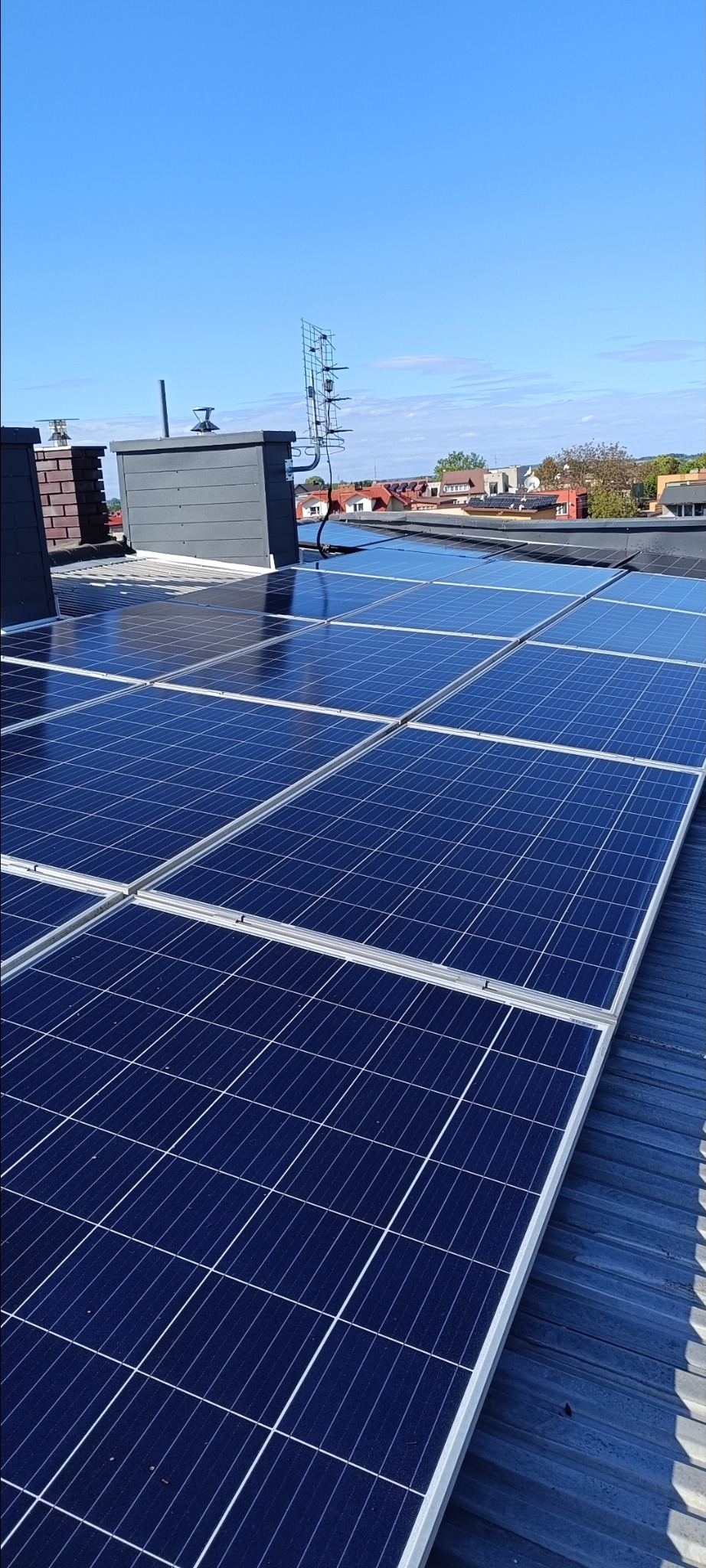 Błękitne panele słoneczne na dachu budynku w słoneczny dzień. Widoczne kominy, antena i fragmenty zabudowy w tle. Perspektywa z dołu, podkreślająca instalację.