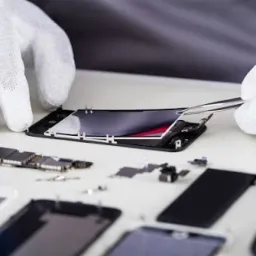 Otwarty smartfon na białym stole, rozłożony na części z widoczną płytą główną, baterią i wyświetlaczem, naprawiany za pomocą pęsety przez osobę w białych rękawiczkach.