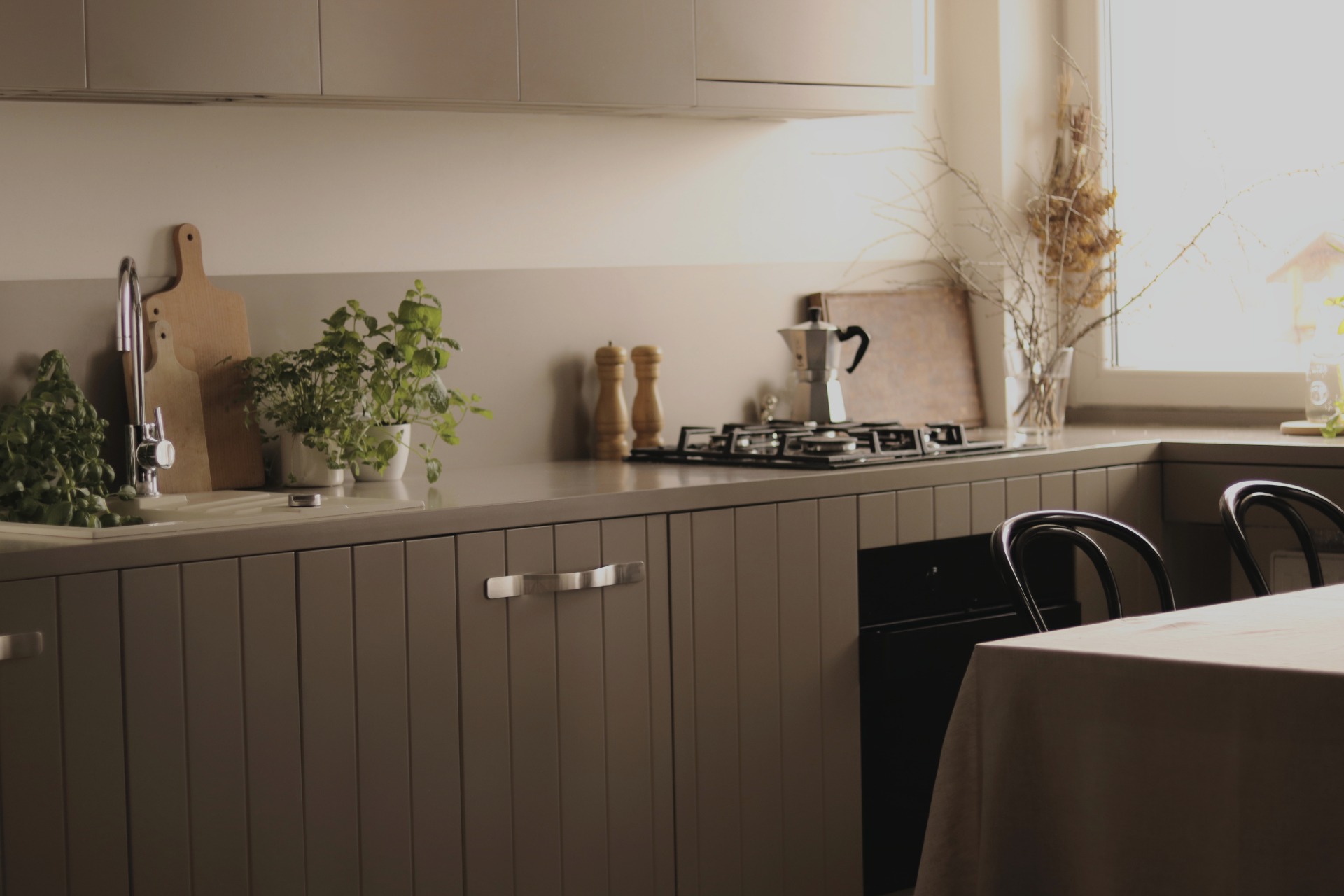 Kuchnia w stonowanej kolorystyce z frontami szafek w pionowe panele, zioła w doniczkach przy zlewie, młynek do kawy na płycie indukcyjnej oraz suszone gałązki w wazonie przy oknie.