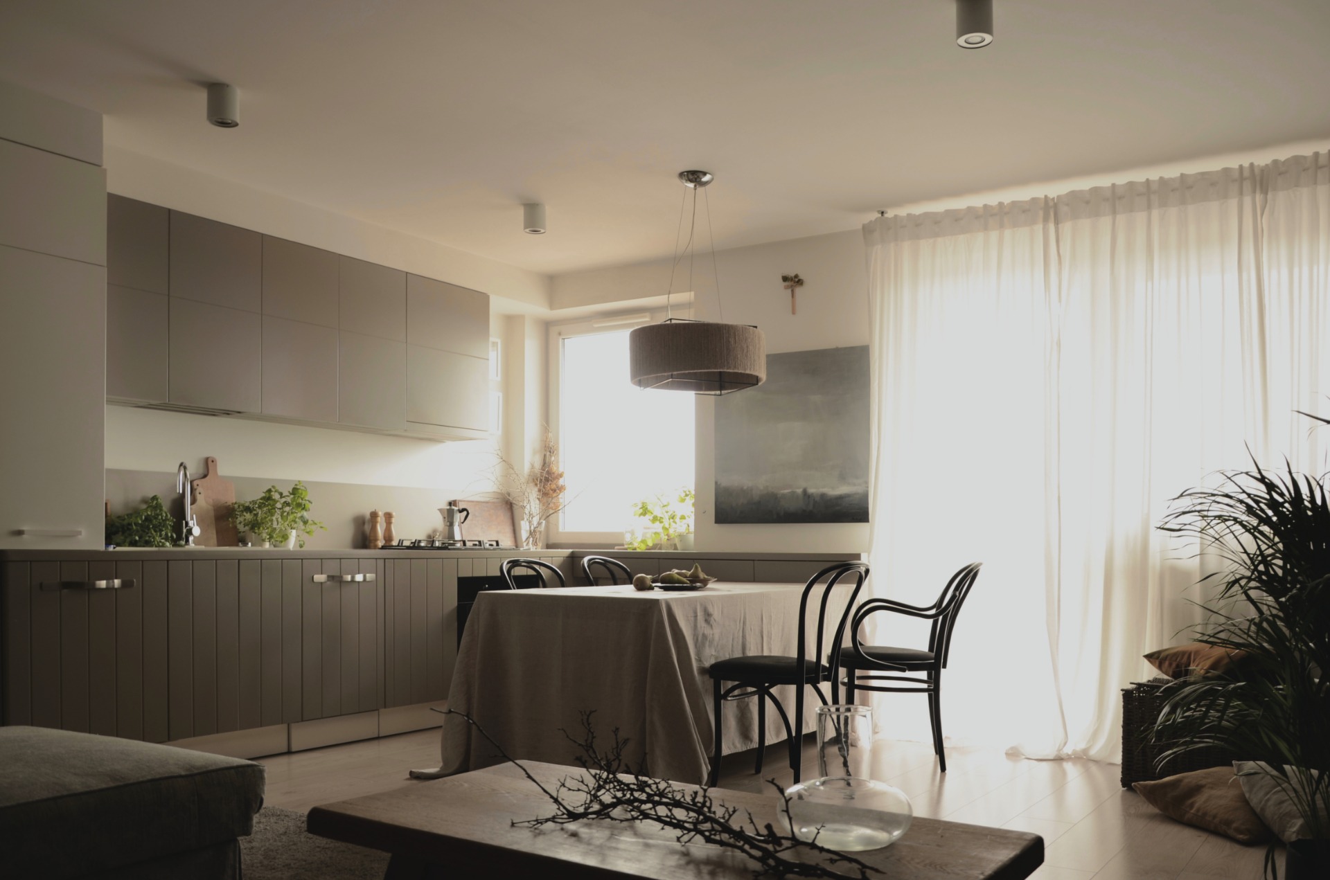 Jasne wnętrze z szarymi szafkami kuchennymi, stołem nakrytym beżowym obrusem, czarnymi krzesłami Thonet i dekoracjami z suszonych gałęzi na drewnianym stole kawowym.